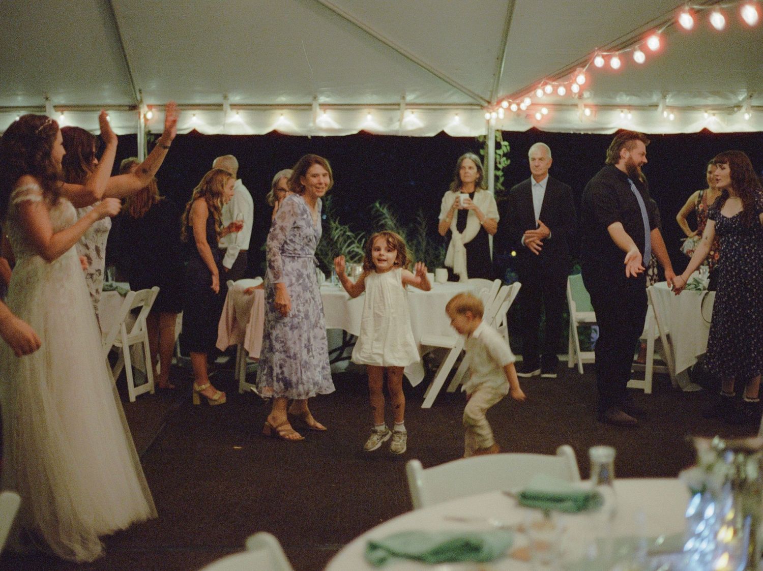 Guests dancing and celebrating under a tent at an outdoor wedding reception with string lights.