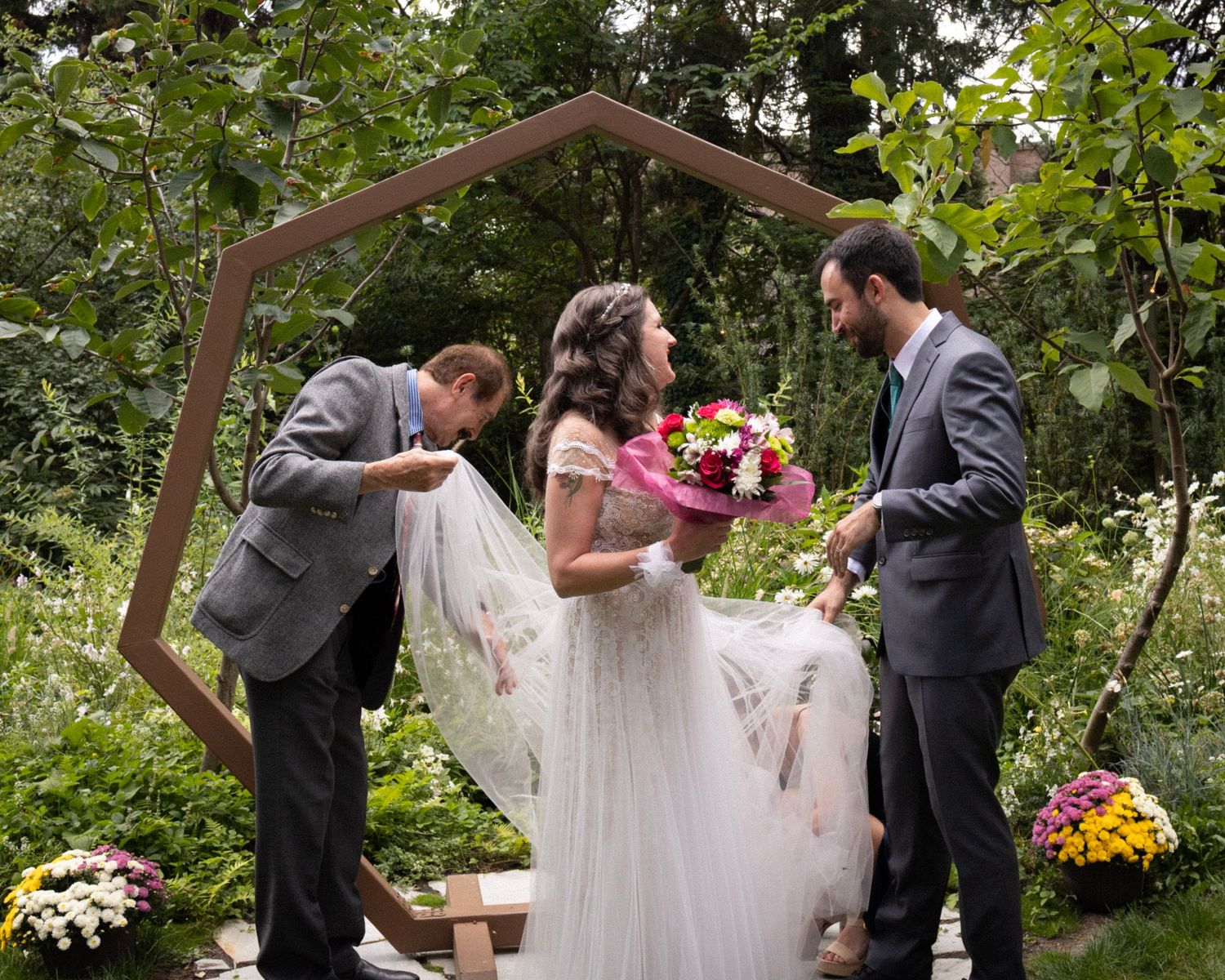 Wedding ceremony moment as officiant lifts bride's veil while groom watches in garden setting.