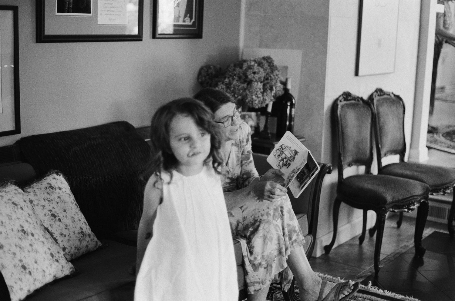 Black and white photo of a young girl in white dress standing with an elderly woman reading a book on a sofa.
