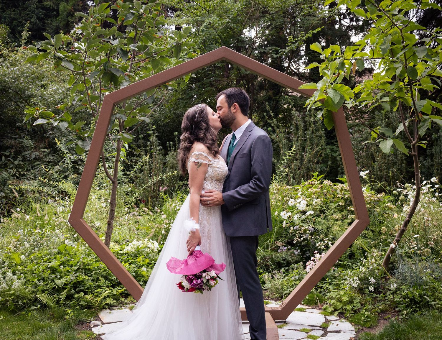 Couple sharing first kiss under wooden geometric arch in lush garden wedding ceremony.