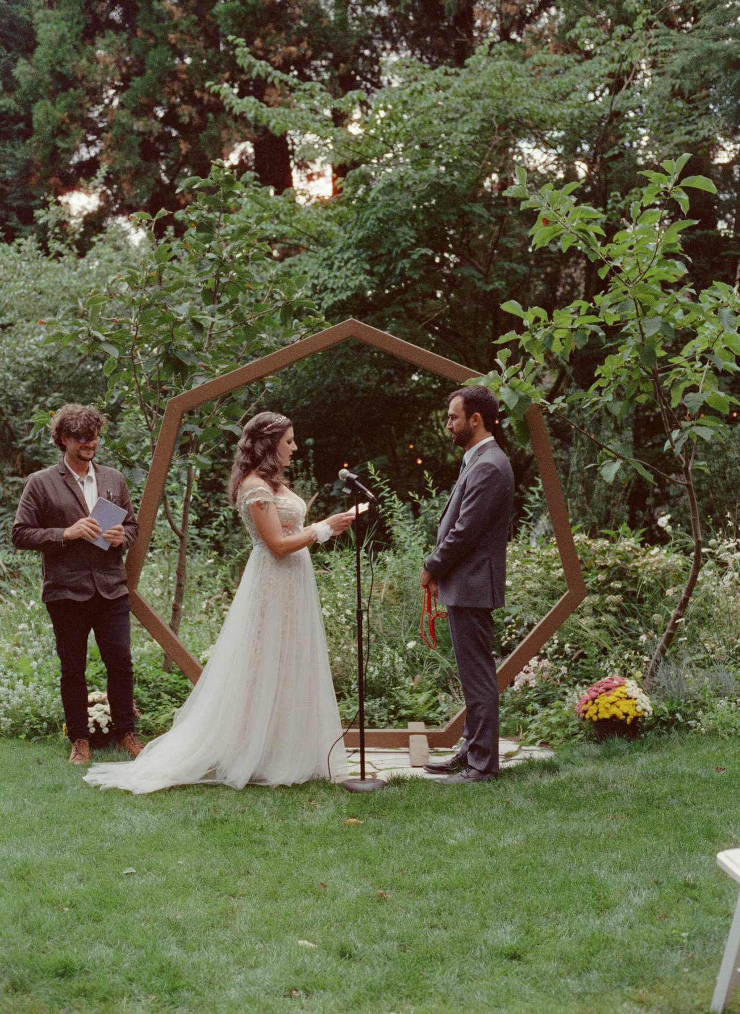 Outdoor wedding ceremony with couple exchanging vows under a geometric wooden arch surrounded by lush greenery.