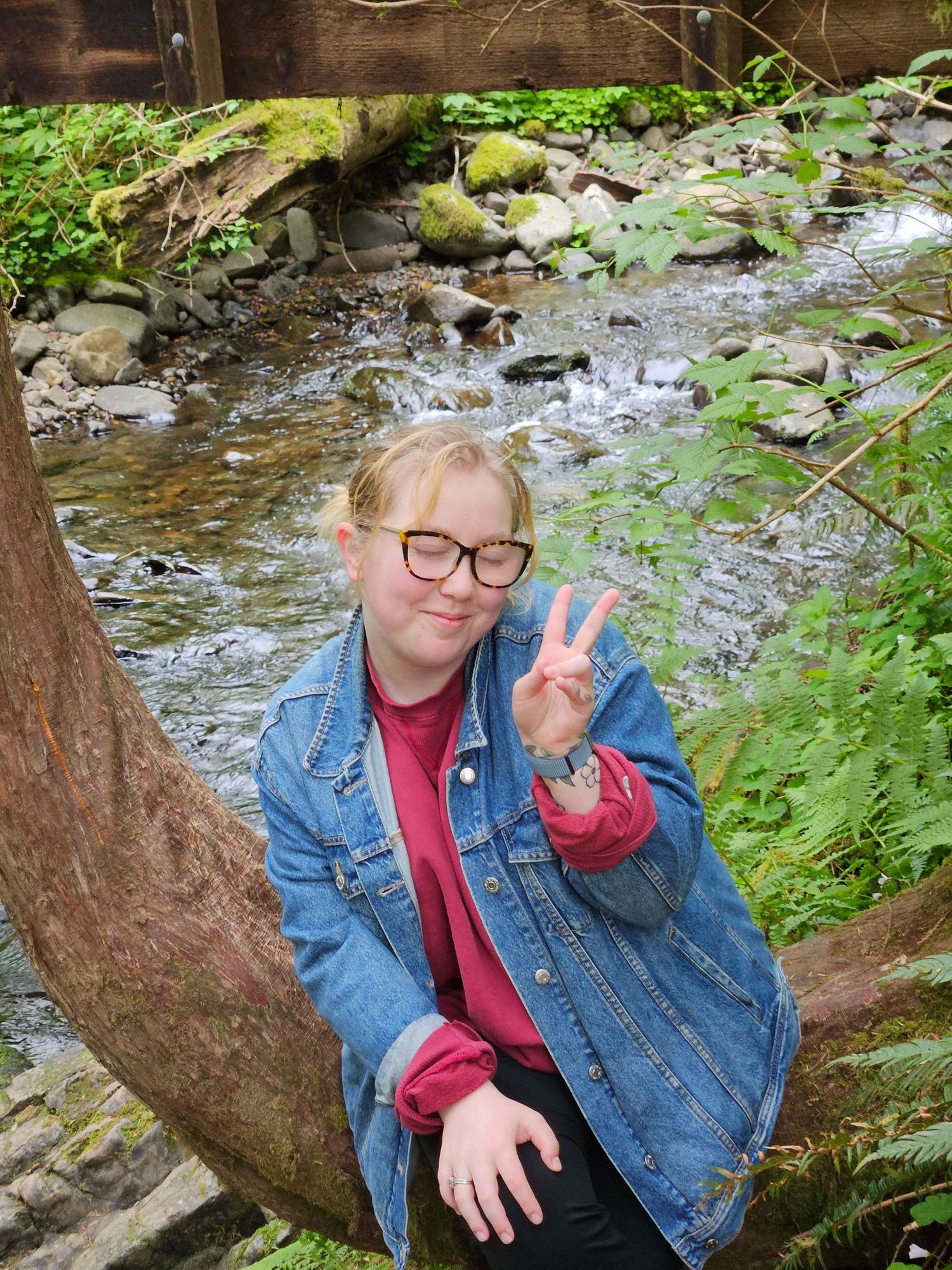 Smiling young woman with glasses flashing peace sign while sitting by a mossy creek in forest.