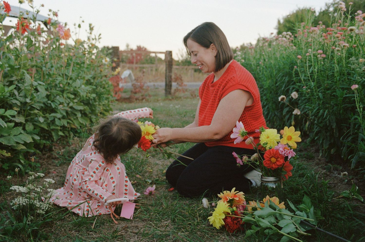 Woman in red top kneels in flower garden sharing colorful blooms with young girl in pink dress on sunny day.