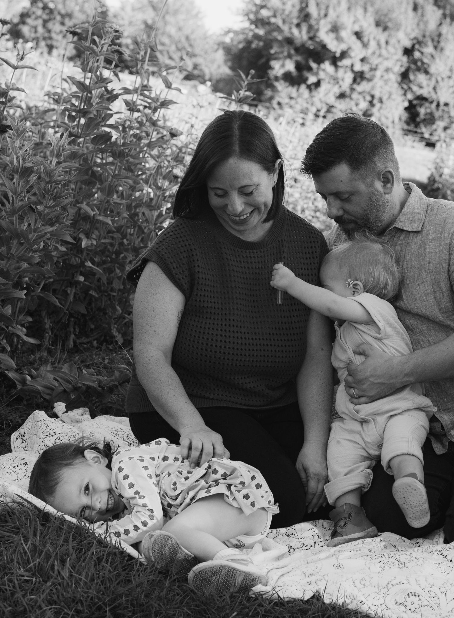 Black and white family photo of parents smiling at two babies on a blanket outdoors among lush greenery.