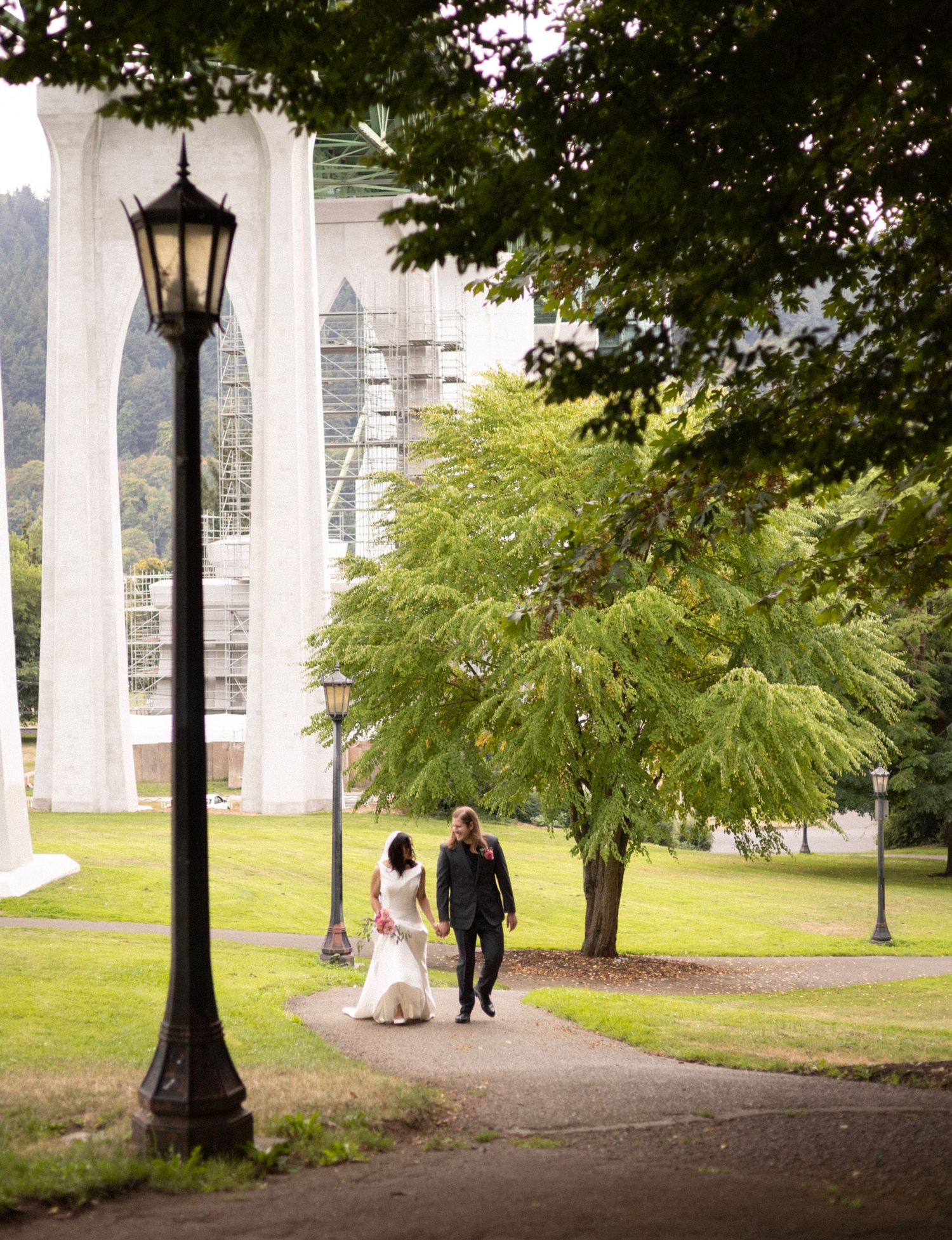 Bride and groom walking on a park path beneath St. Johns Bridge arches, surrounded by lush green trees.