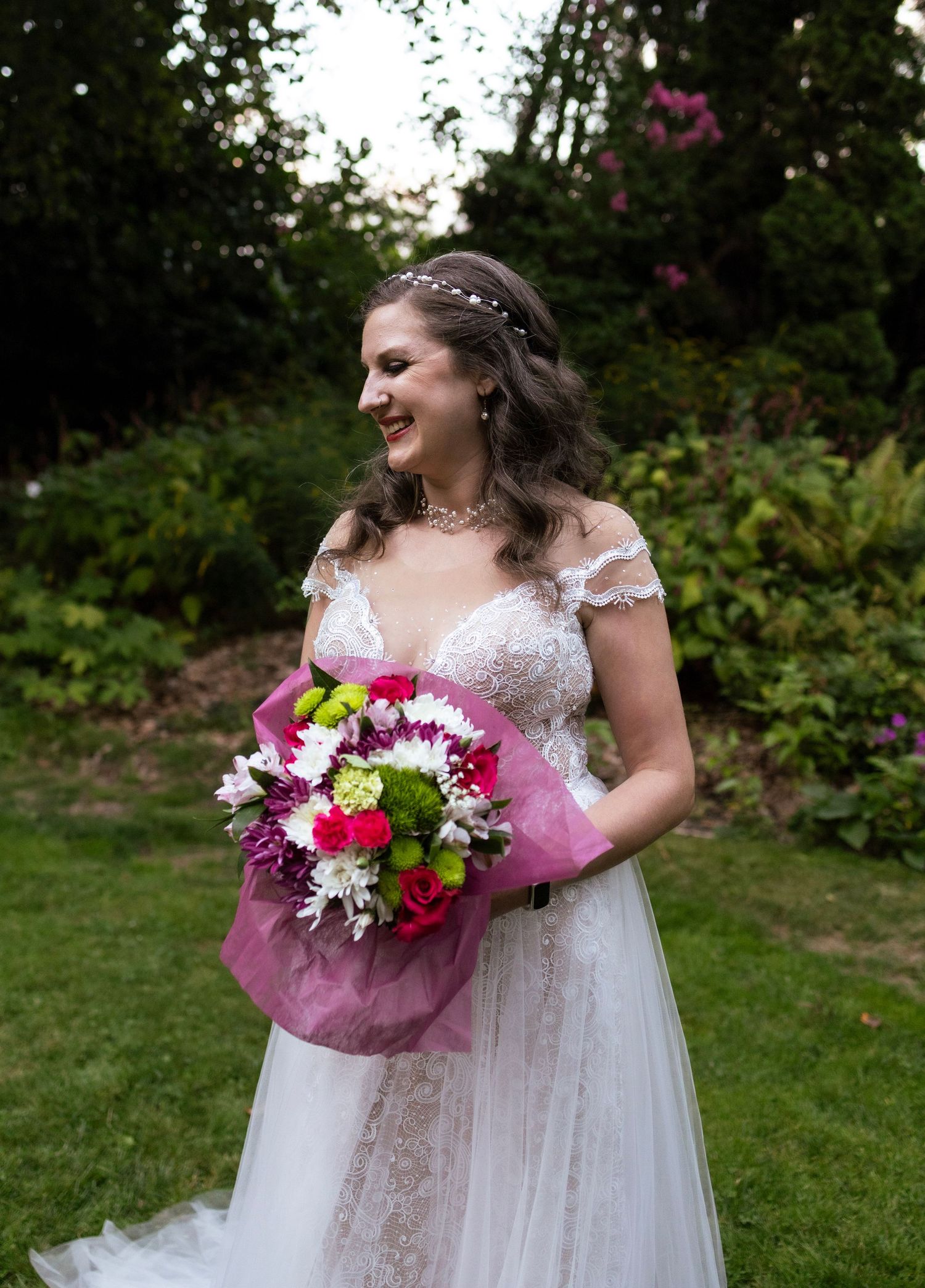 Smiling bride in a lace off-shoulder gown holding a vibrant pink and green bouquet in a garden.