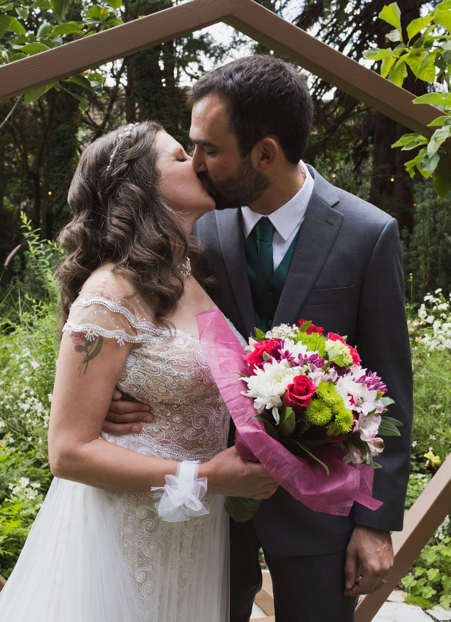 Bride and groom sharing a first kiss under a wooden arch while holding a colorful wedding bouquet.