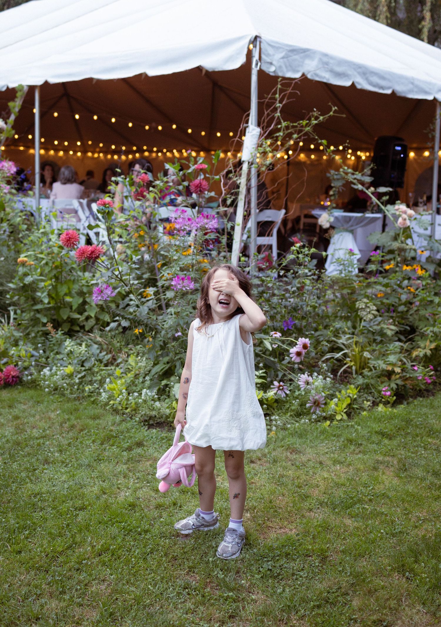A laughing little girl in a white dress holds a pink stuffed animal near a floral garden at a wedding.