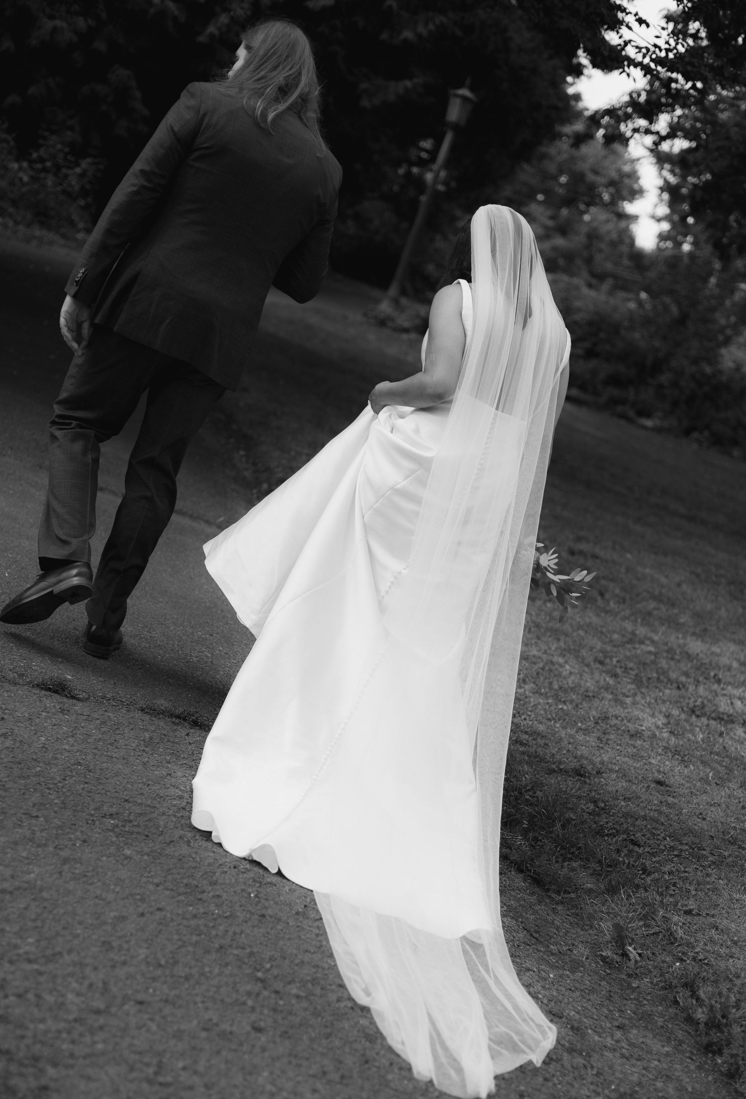 Black and white photo of bride in flowing veil and gown walking with groom in park.