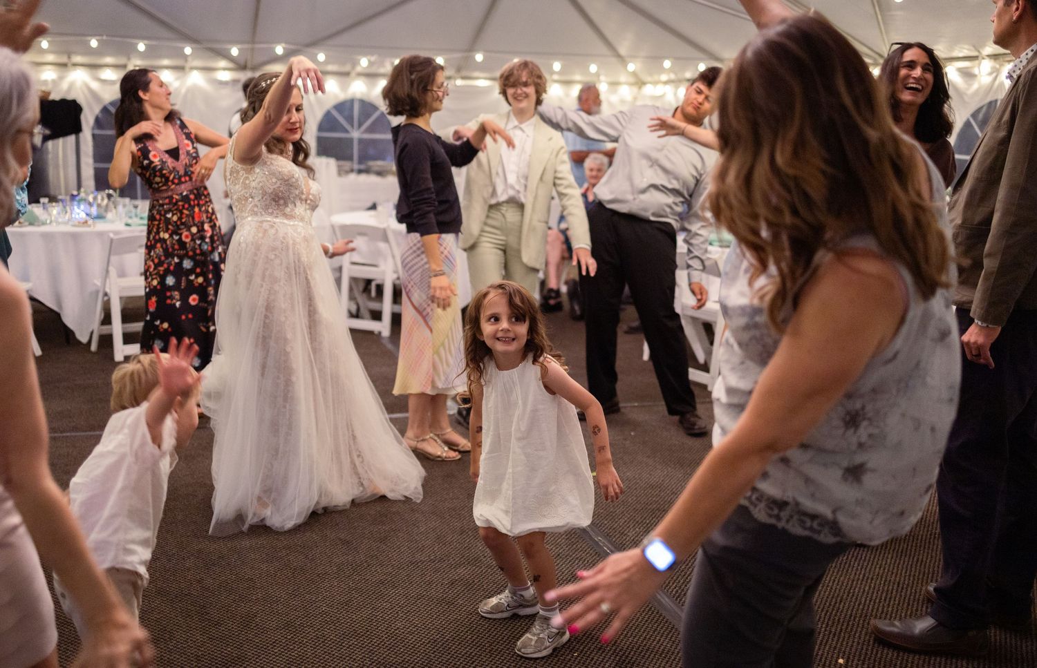 Young girl smiles on dance floor surrounded by wedding guests dancing under tent.