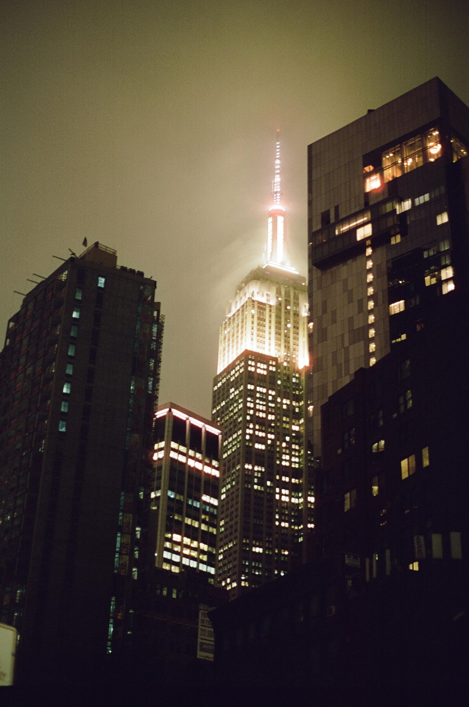 The Empire State Building illuminated at night, partially shrouded in fog, surrounded by glowing Manhattan skyscrapers.