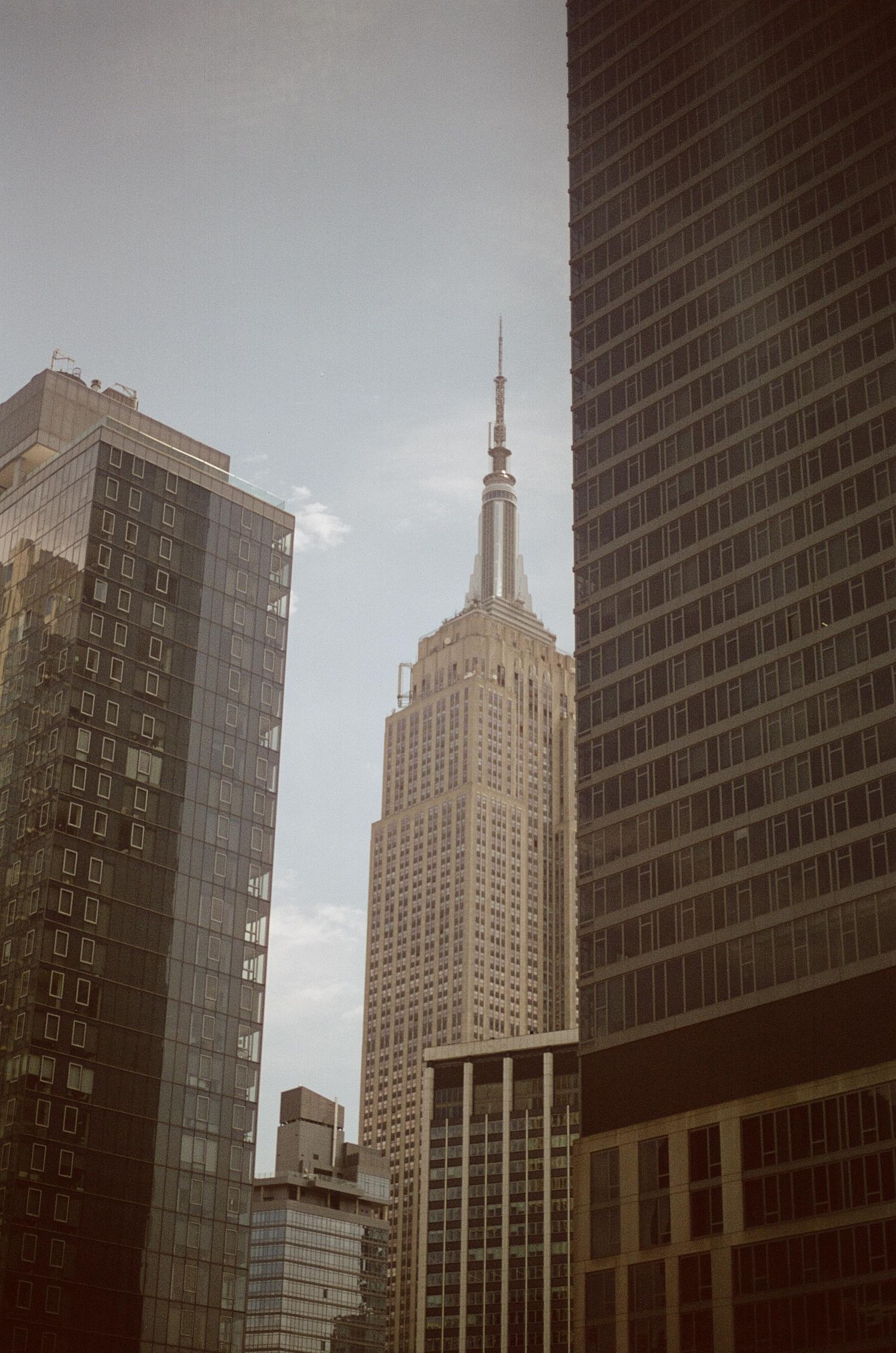 The iconic Empire State Building rises between modern glass skyscrapers in Midtown Manhattan under a clear blue sky.