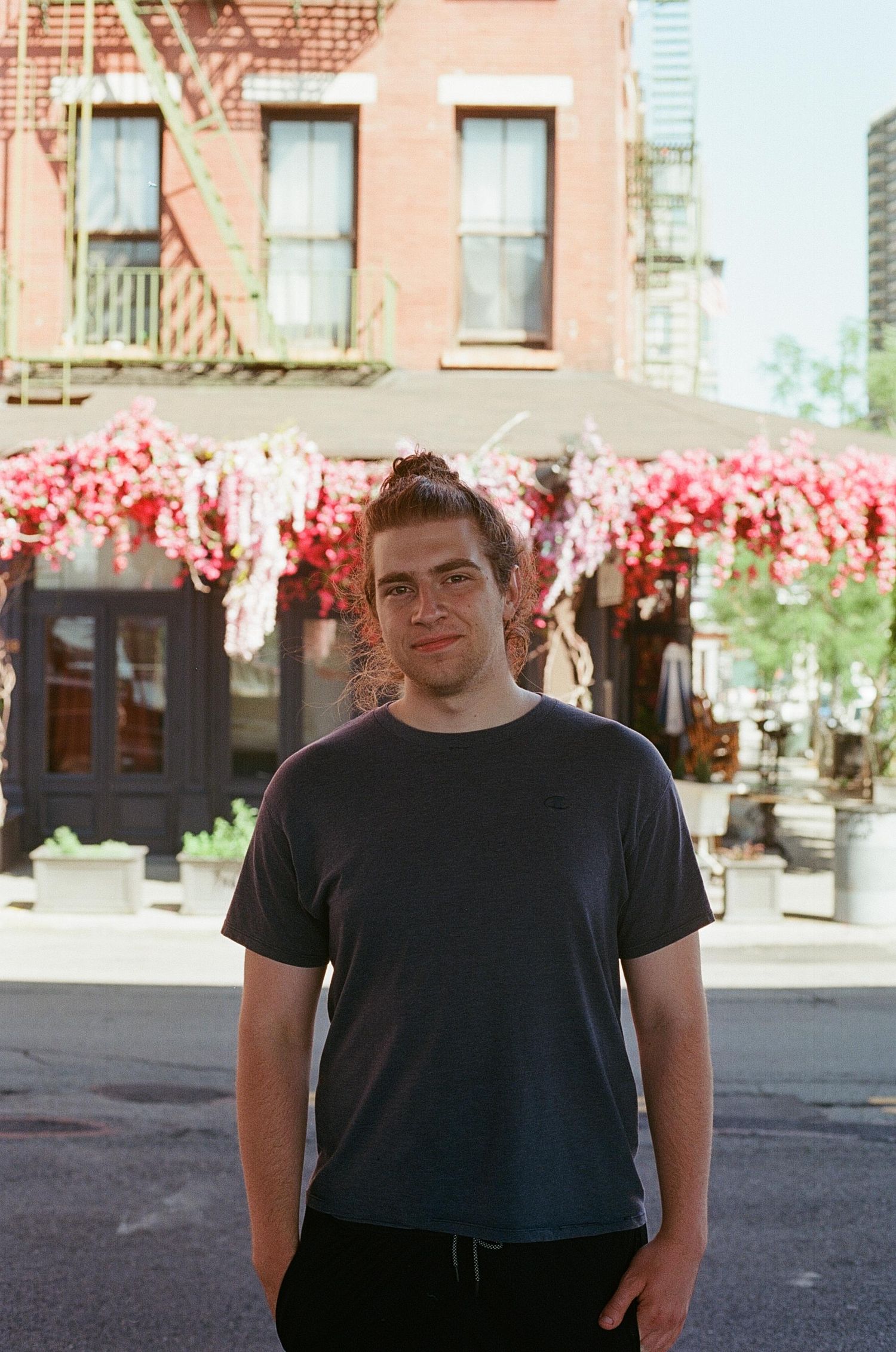 Young man in a navy t-shirt standing on a sunny New York City street with pink floral decorations behind him.