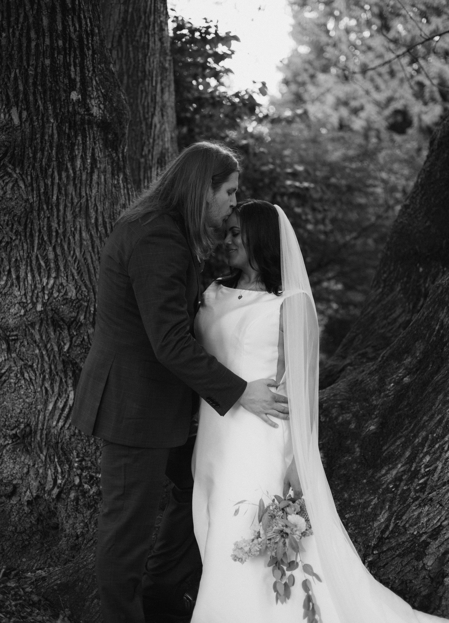 Black and white wedding photo of bride in veil holding bouquet and groom embracing between large trees outdoors.