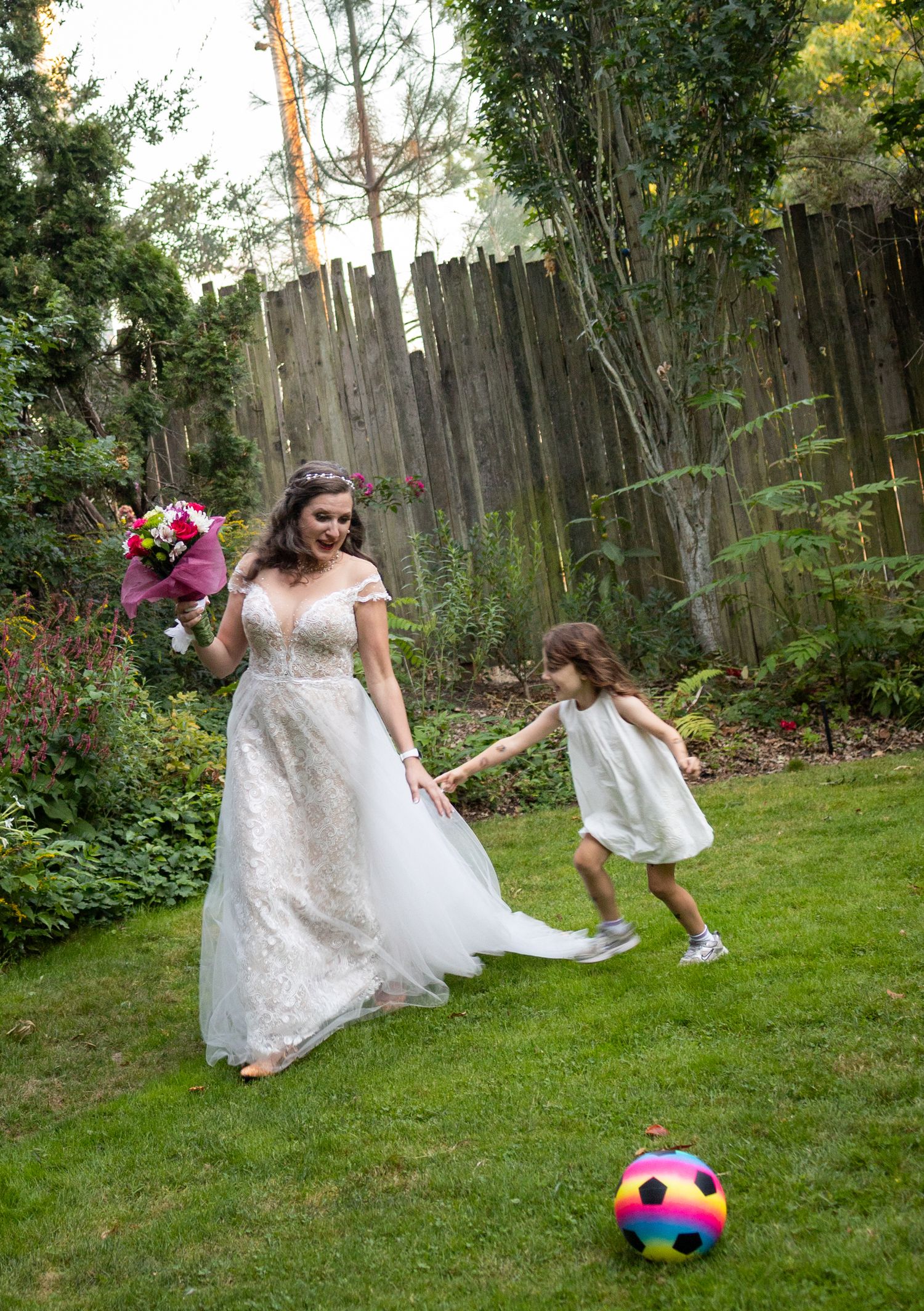 A bride in a lace gown holds a bouquet while playing with a young flower girl on a garden lawn.
