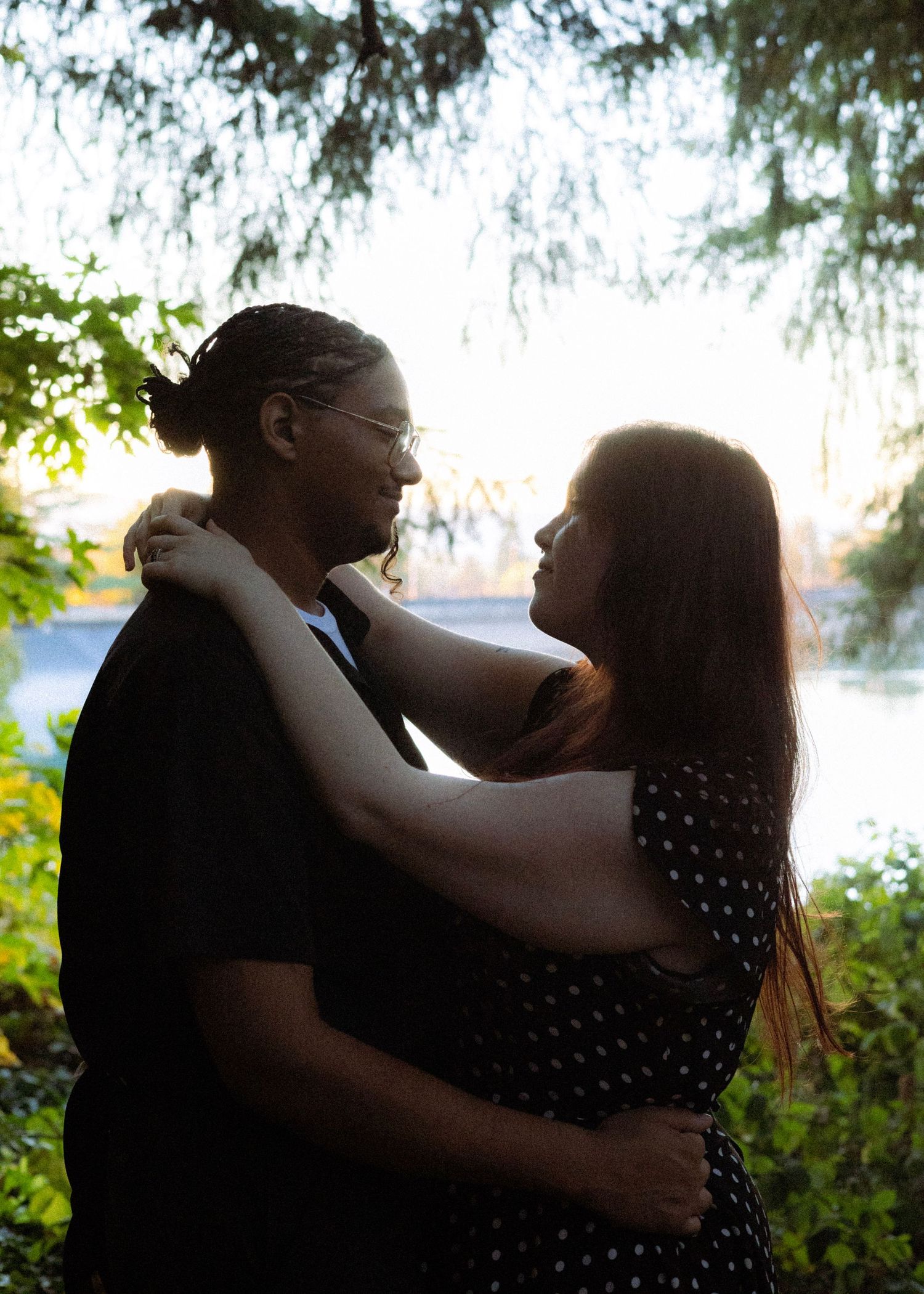 Couple embraces at golden hour near a lake, surrounded by lush green trees in a romantic outdoor setting.