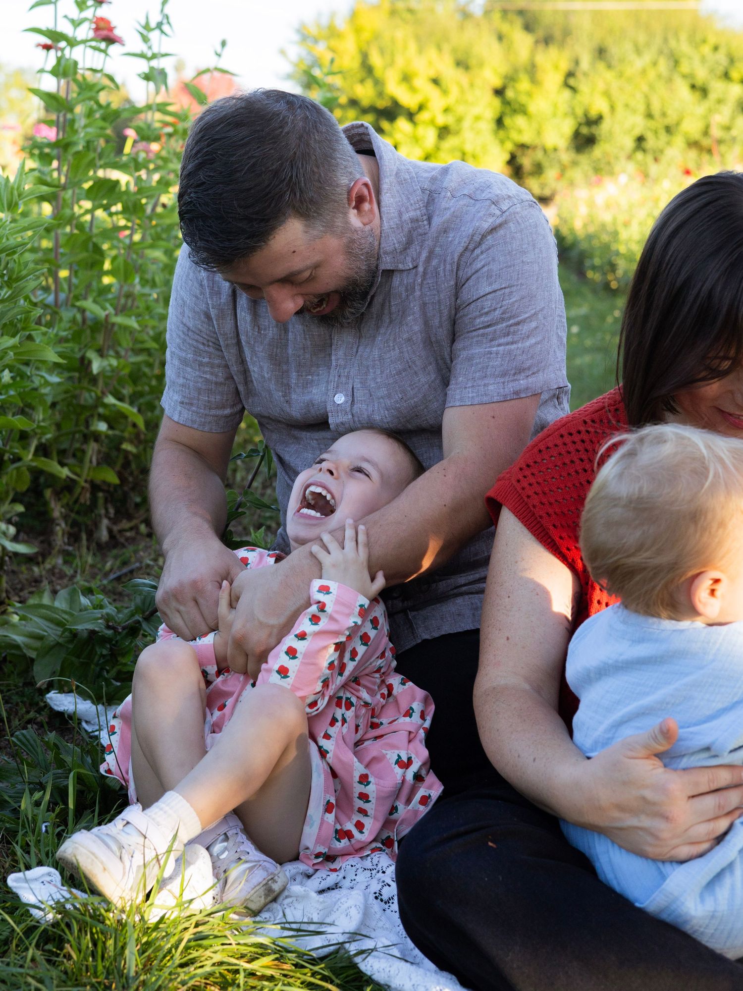 A man tickles a laughing baby girl outdoors while a woman holds a toddler in a sunny garden setting.