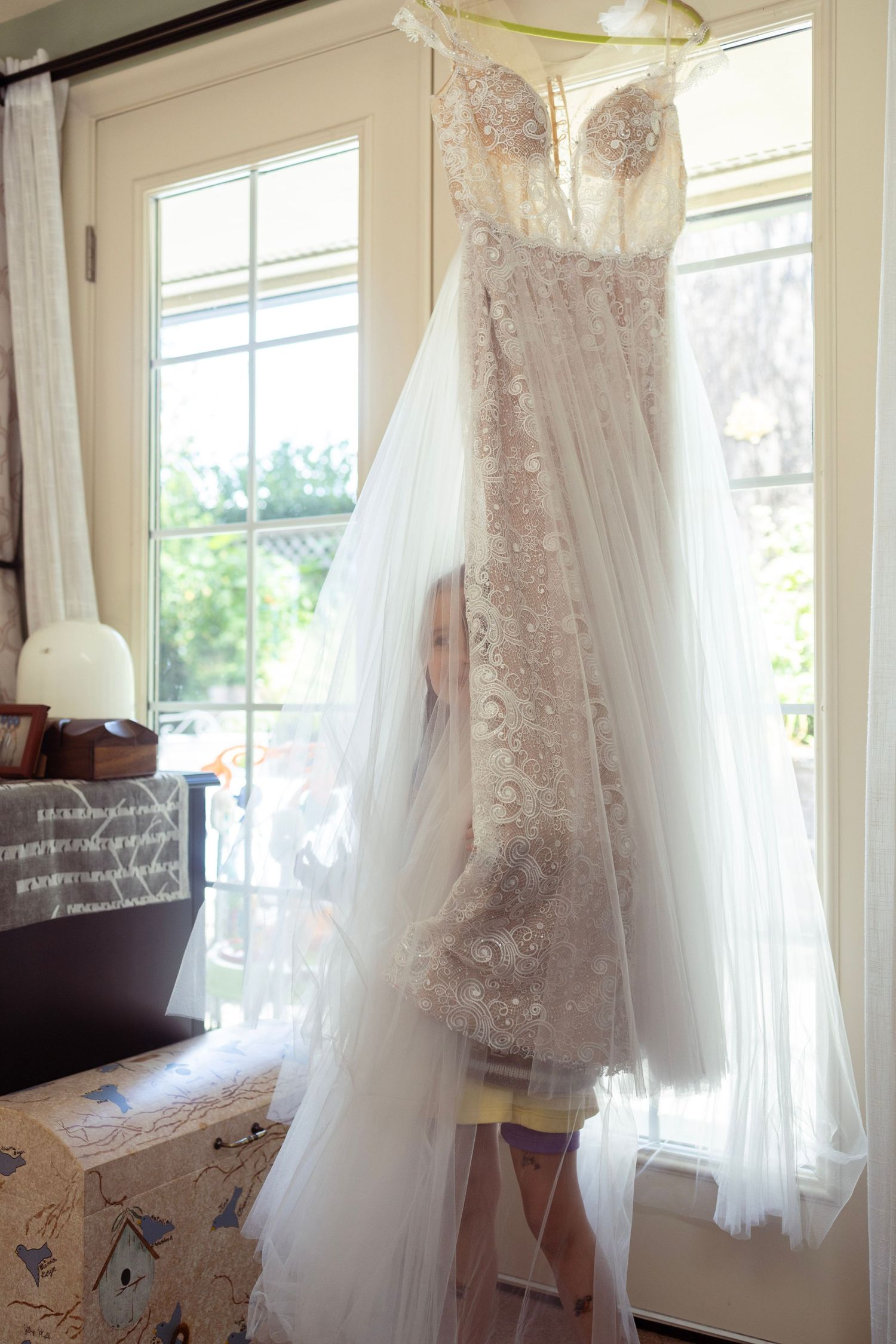 Young flower girl peeking through bridal gown veil hanging in sunlit room before wedding.