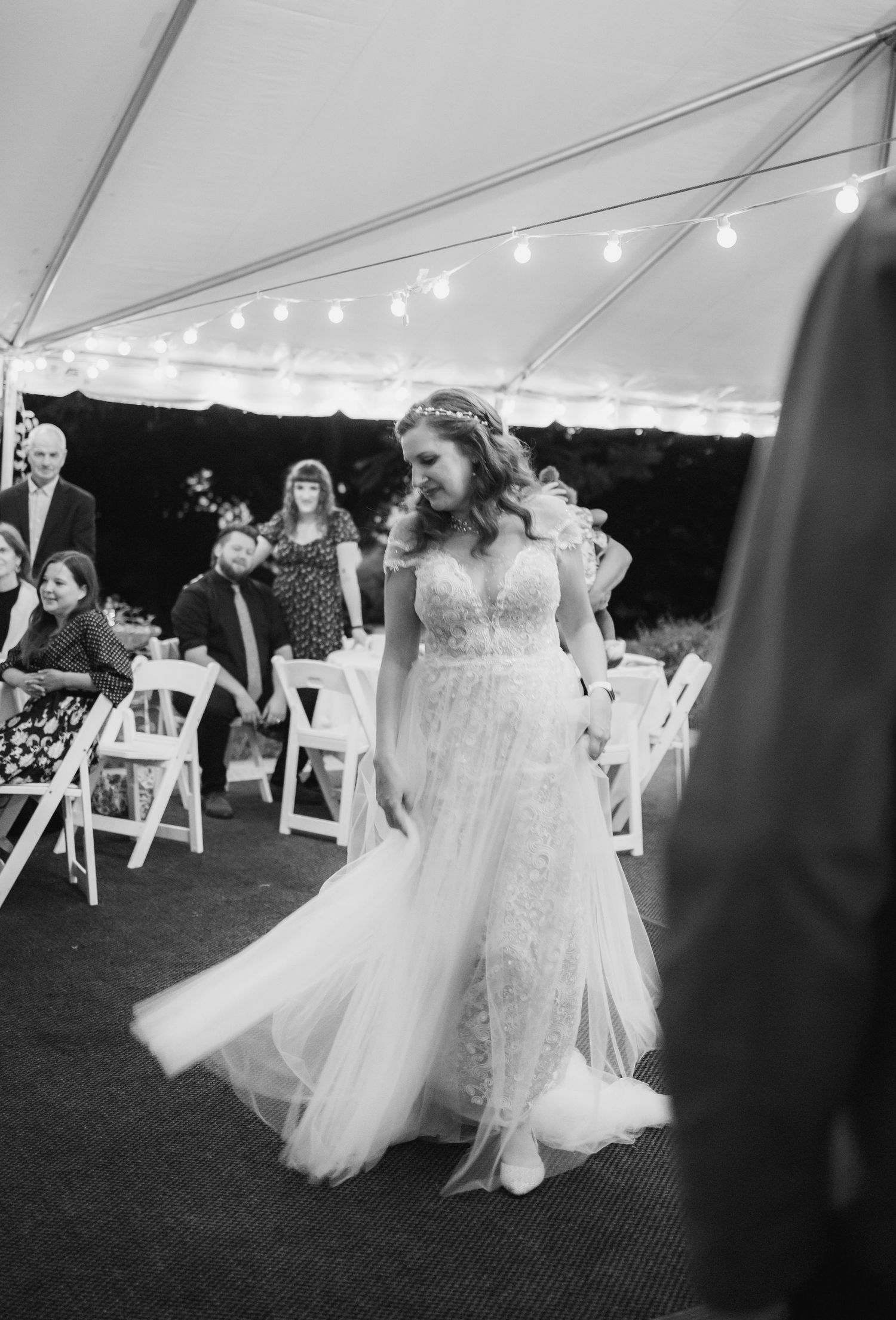 Bride in lace wedding gown walks down aisle under string lights at outdoor tented wedding ceremony.