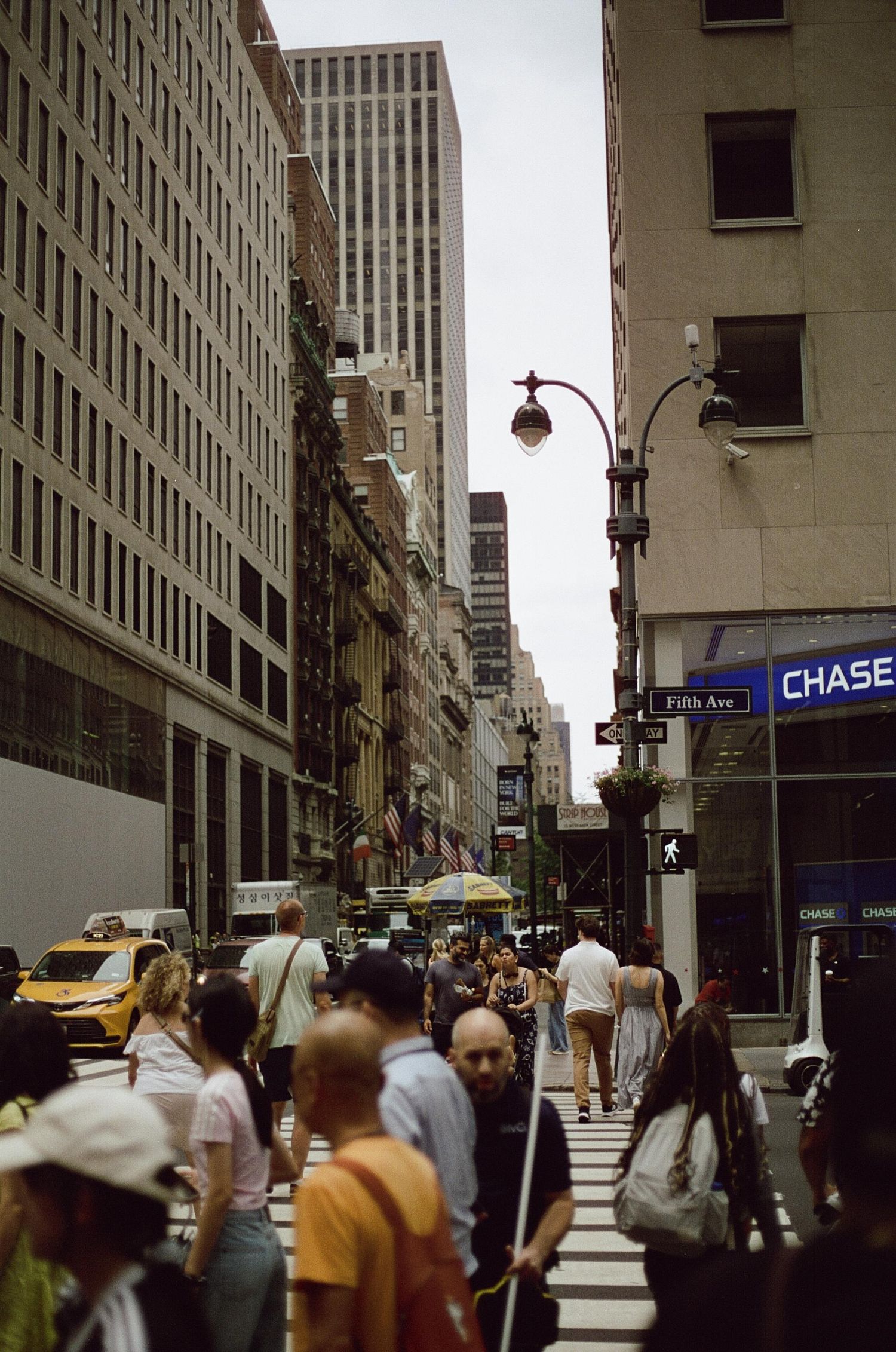 Crowds of pedestrians cross a busy intersection on Fifth Avenue in Midtown Manhattan with a Chase Bank visible.