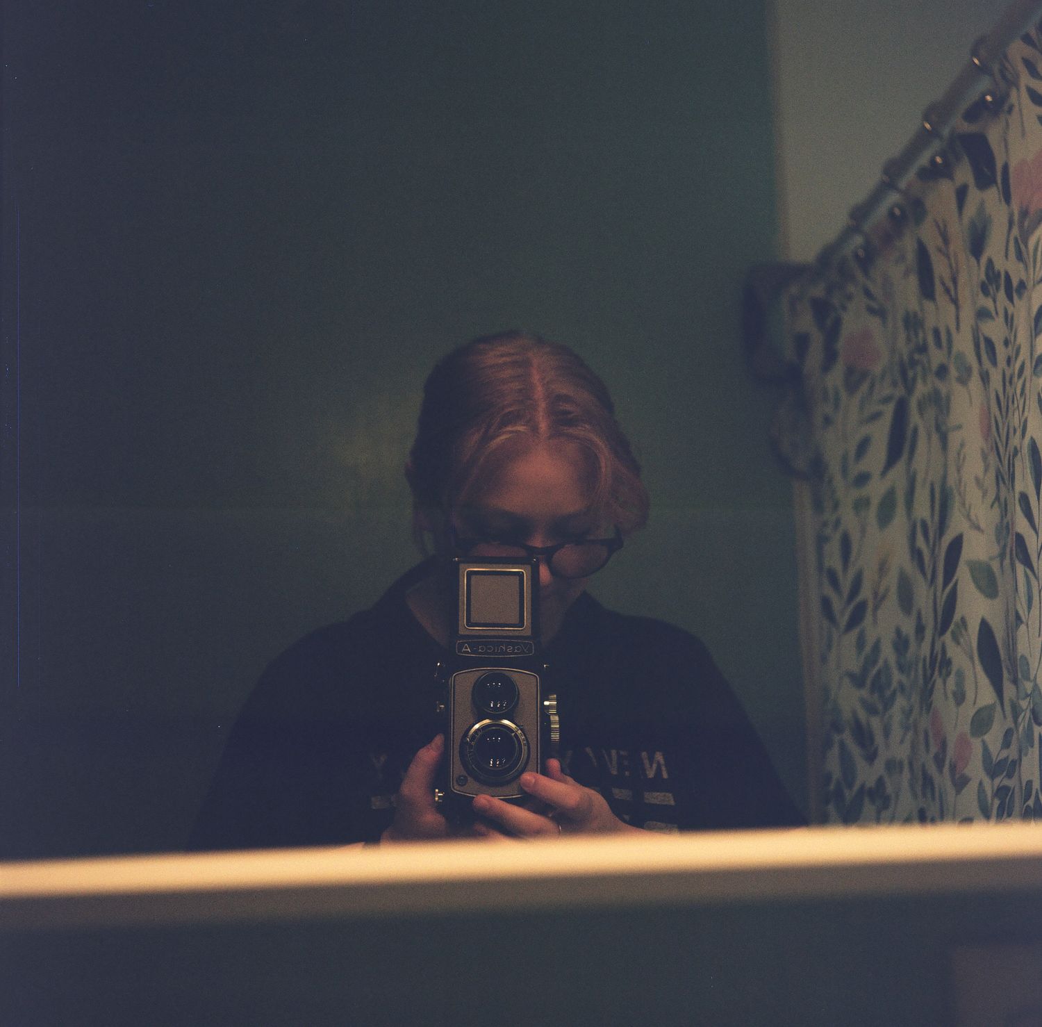 Person taking a mirror selfie in a bathroom with a vintage twin-lens reflex camera.