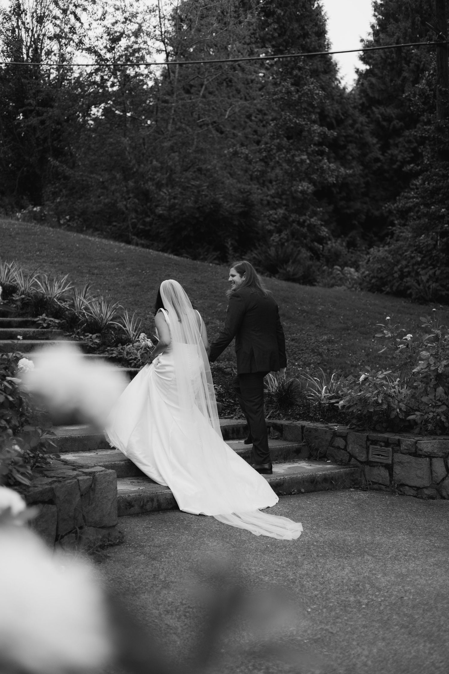 Black and white photo of a bride and groom holding hands walking up stone steps in a lush garden.