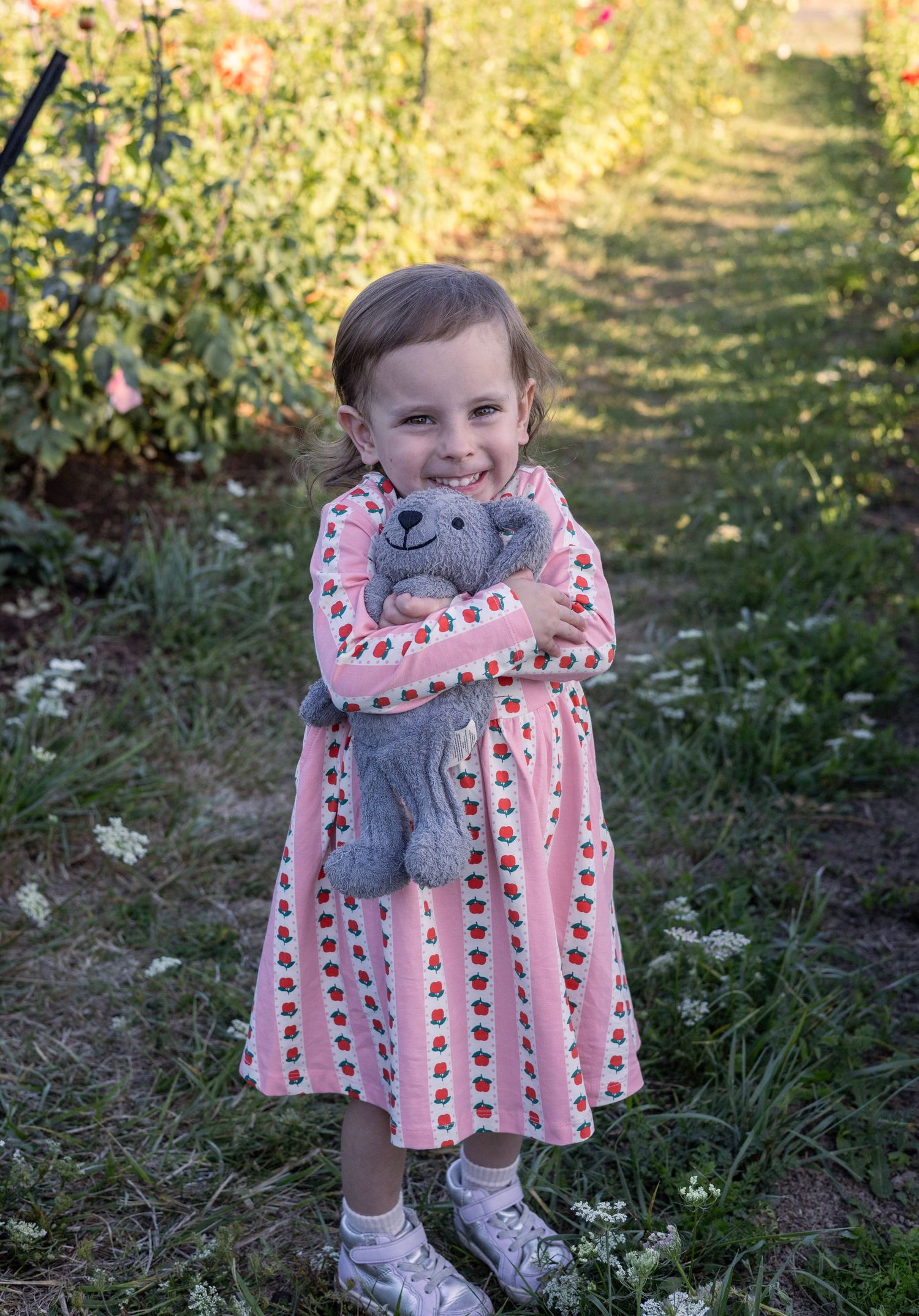 A smiling young girl in a pink strawberry dress hugs a gray teddy bear while standing in a lush garden path.