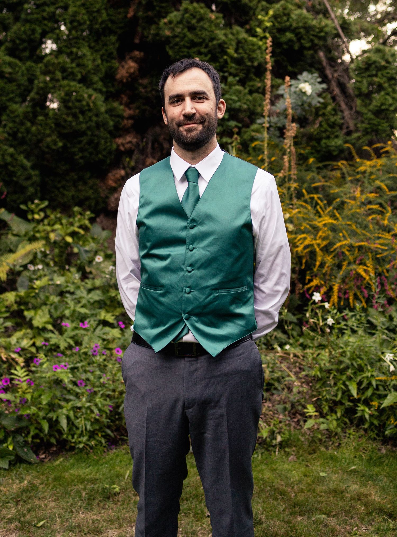 Groom smiles in green vest and tie standing in garden before wedding ceremony.