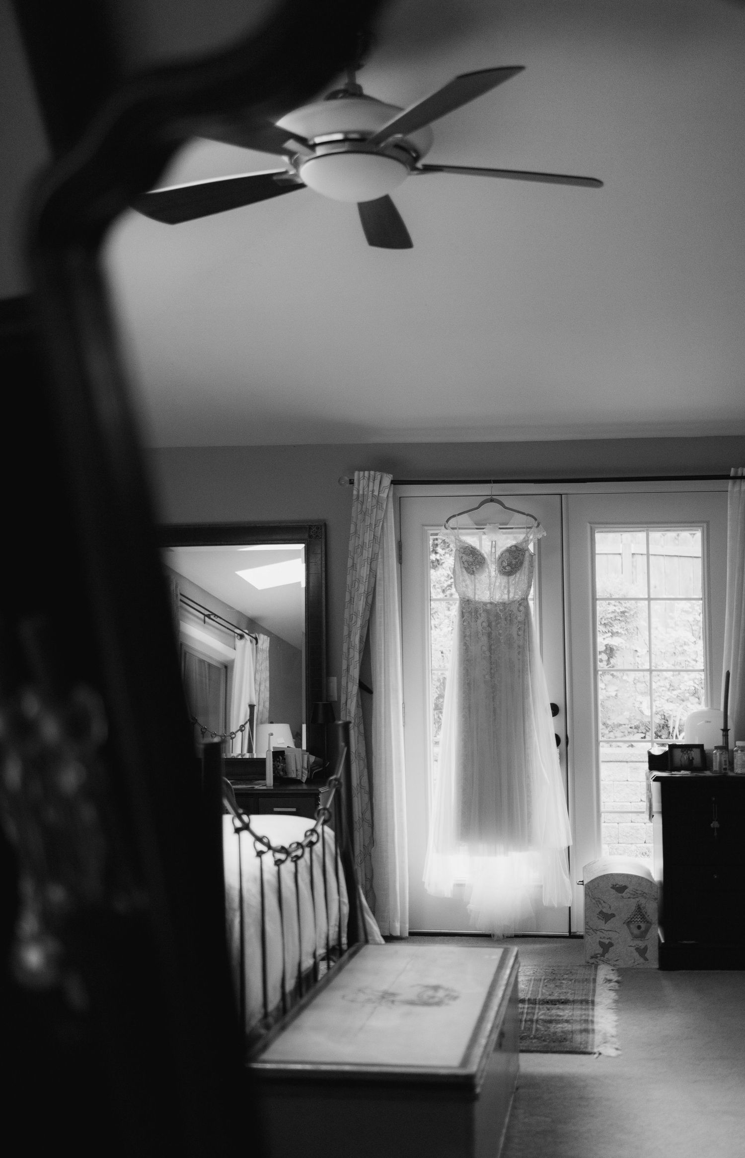A black and white photo of a wedding dress hanging in a sunlit bedroom doorway before the ceremony.