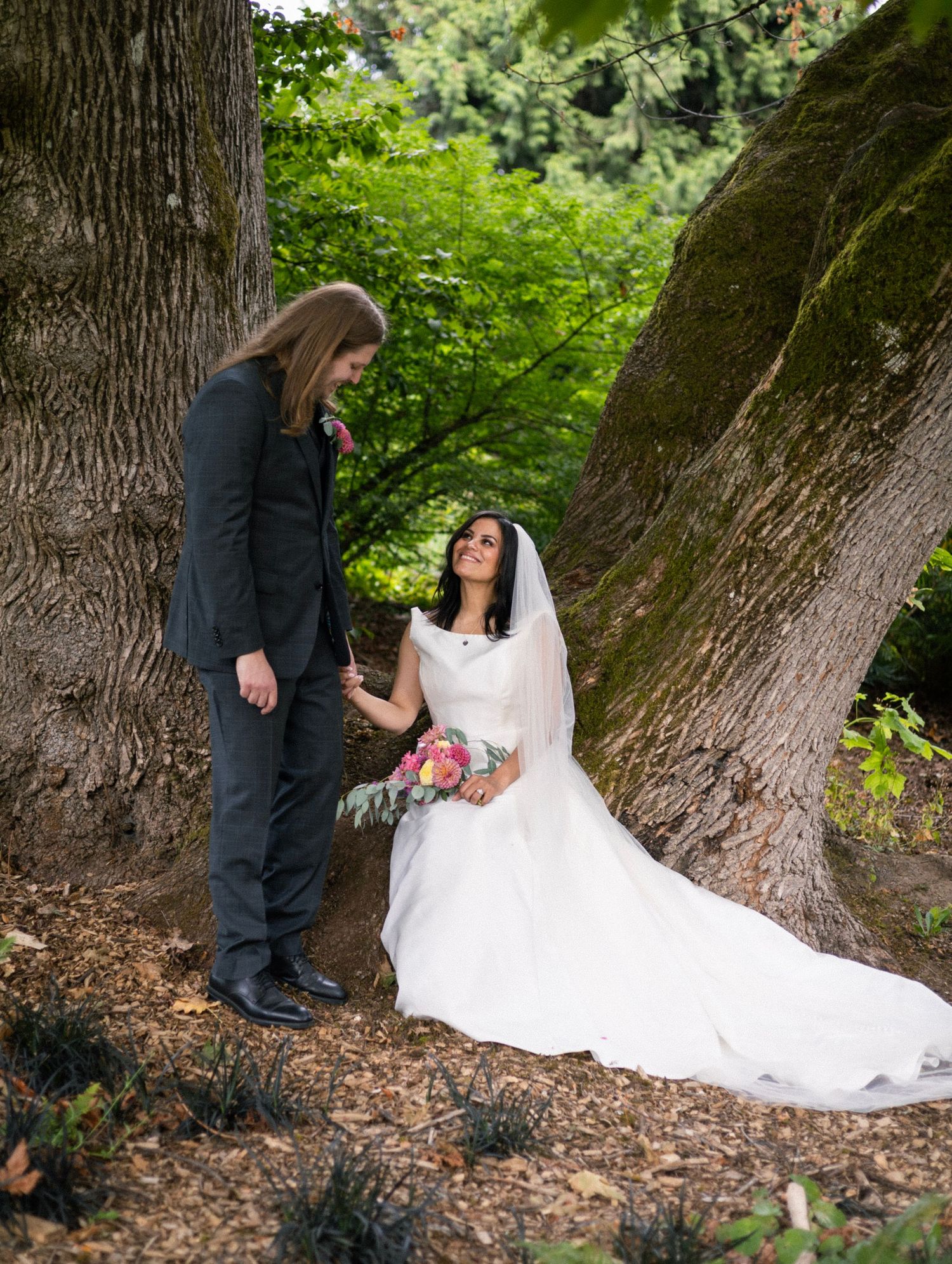 Bride in white gown sits by mossy trees holding bouquet while groom in dark suit looks down at her in forest setting.