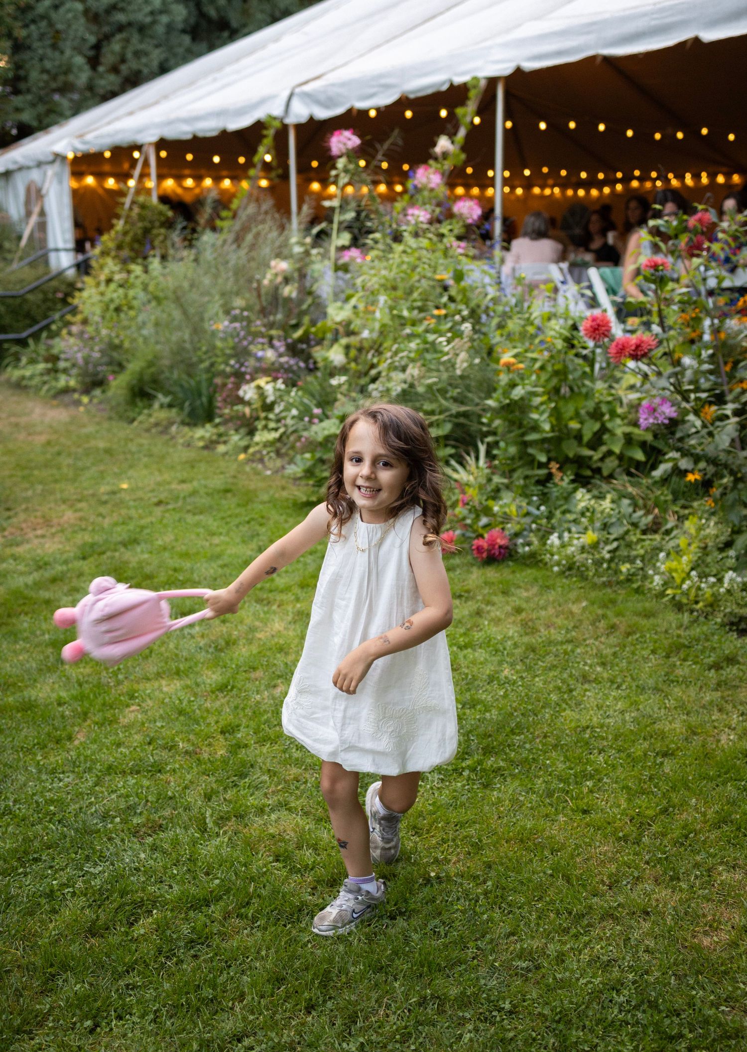 A cheerful little girl in a white dress runs on grass holding a pink toy near a flower garden at a wedding.