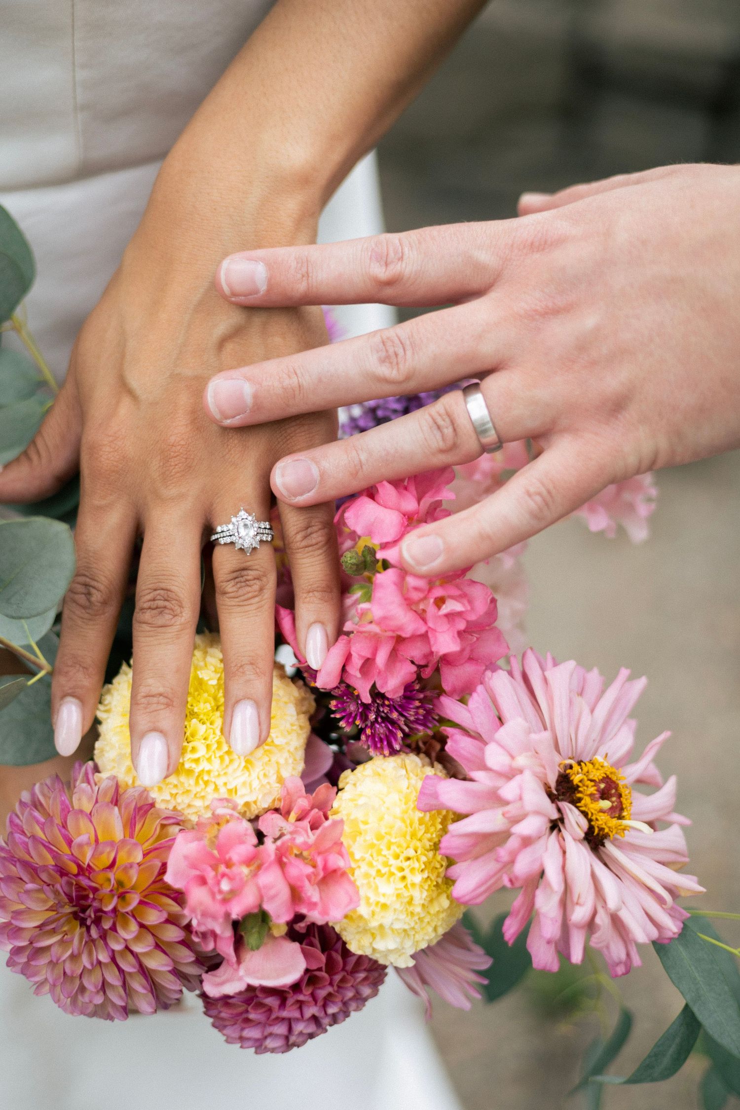 Newlyweds displaying wedding rings on colorful bridal bouquet with pink dahlias and yellow flowers.