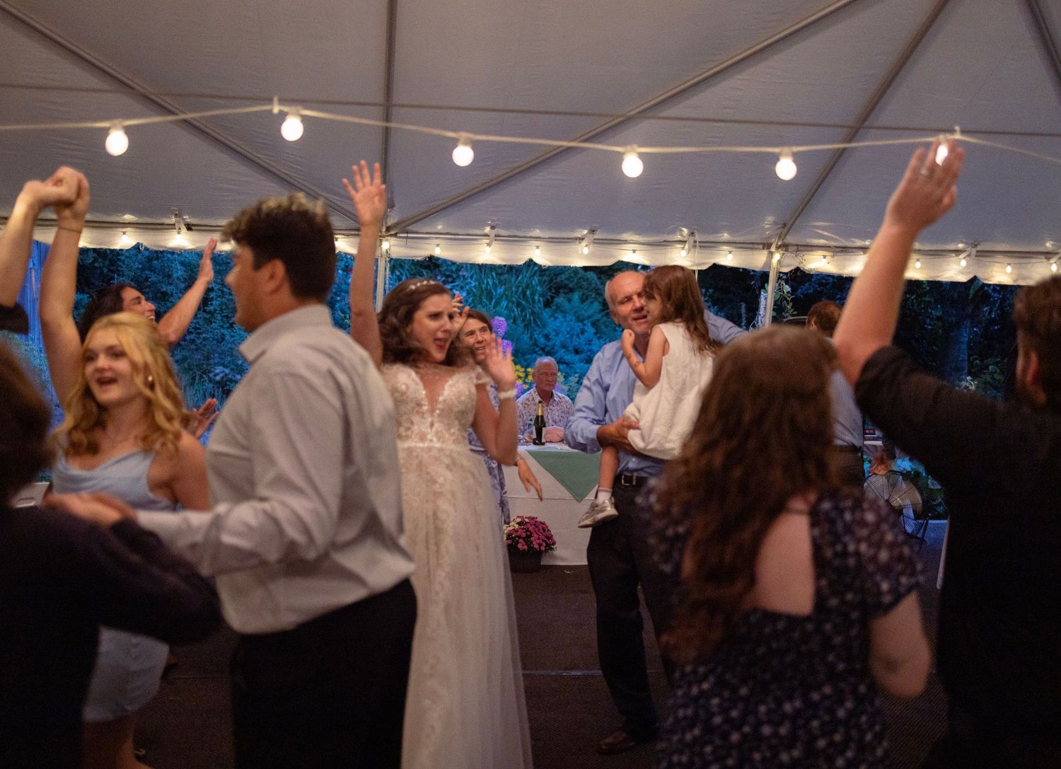 Joyful wedding reception dance floor scene under a tent with bride in white gown dancing among celebrating guests.