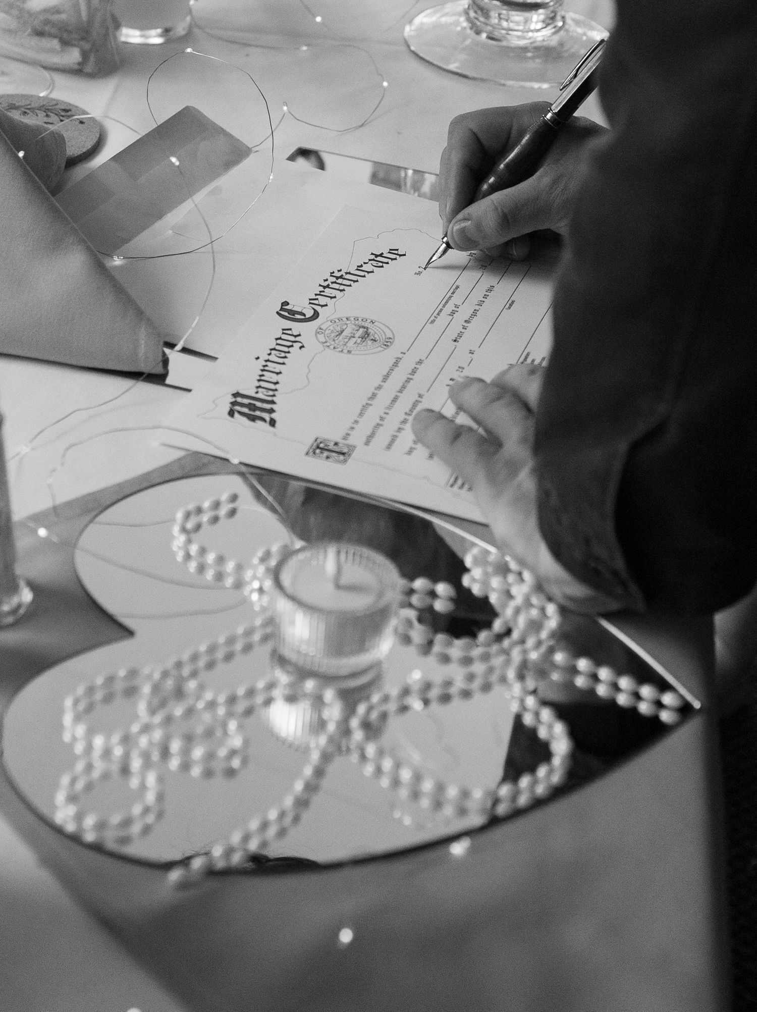 Black and white close-up of a hand signing a marriage certificate on a decorated reception table.