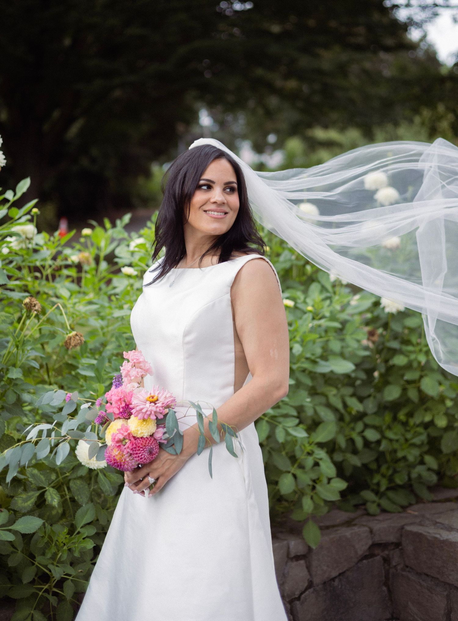 Bride in white sleeveless gown holding pink bouquet with veil flowing in wind among lush garden greenery.