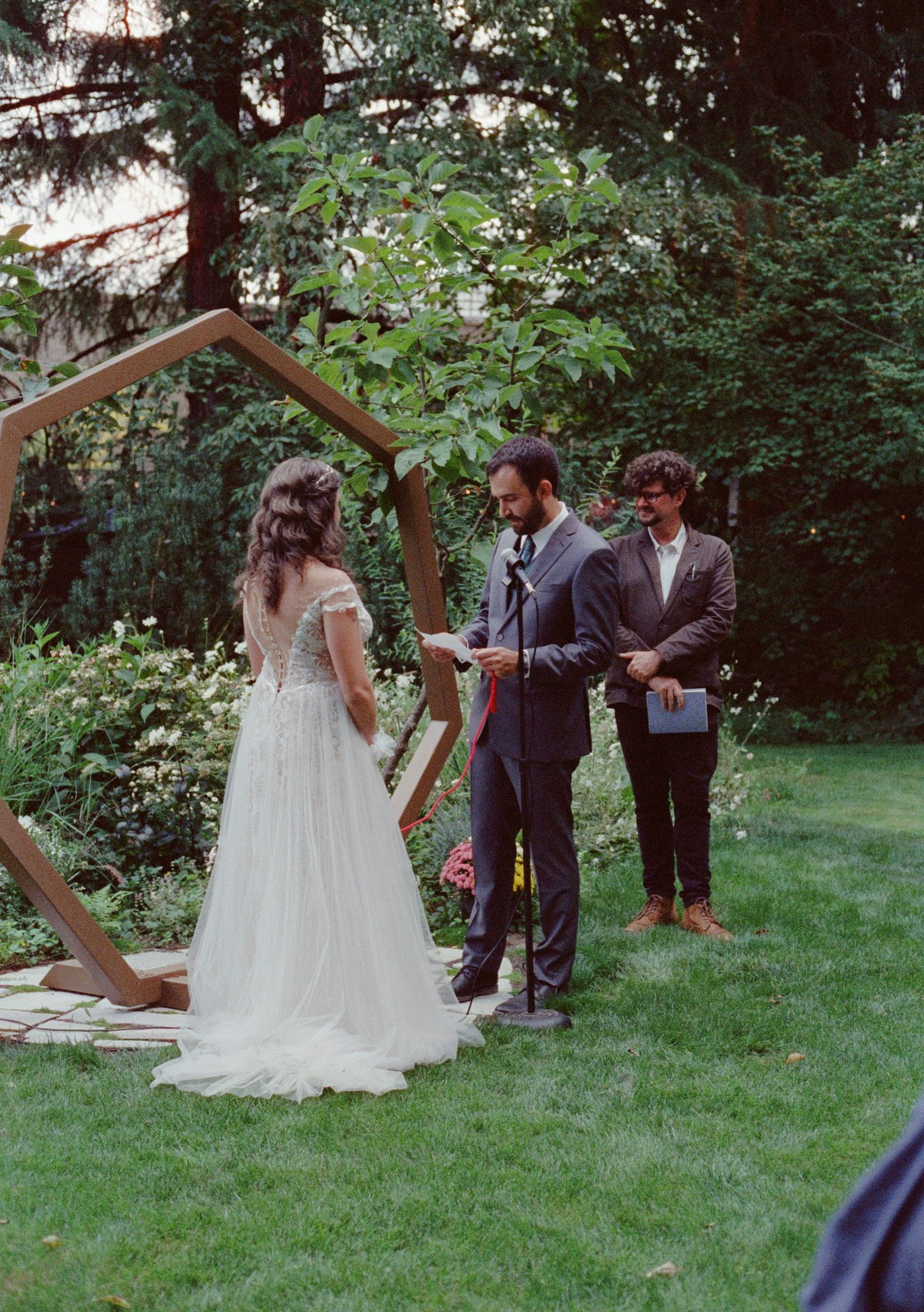 Outdoor garden wedding ceremony with bride in white gown, groom in suit, and officiant under a geometric wooden arch.