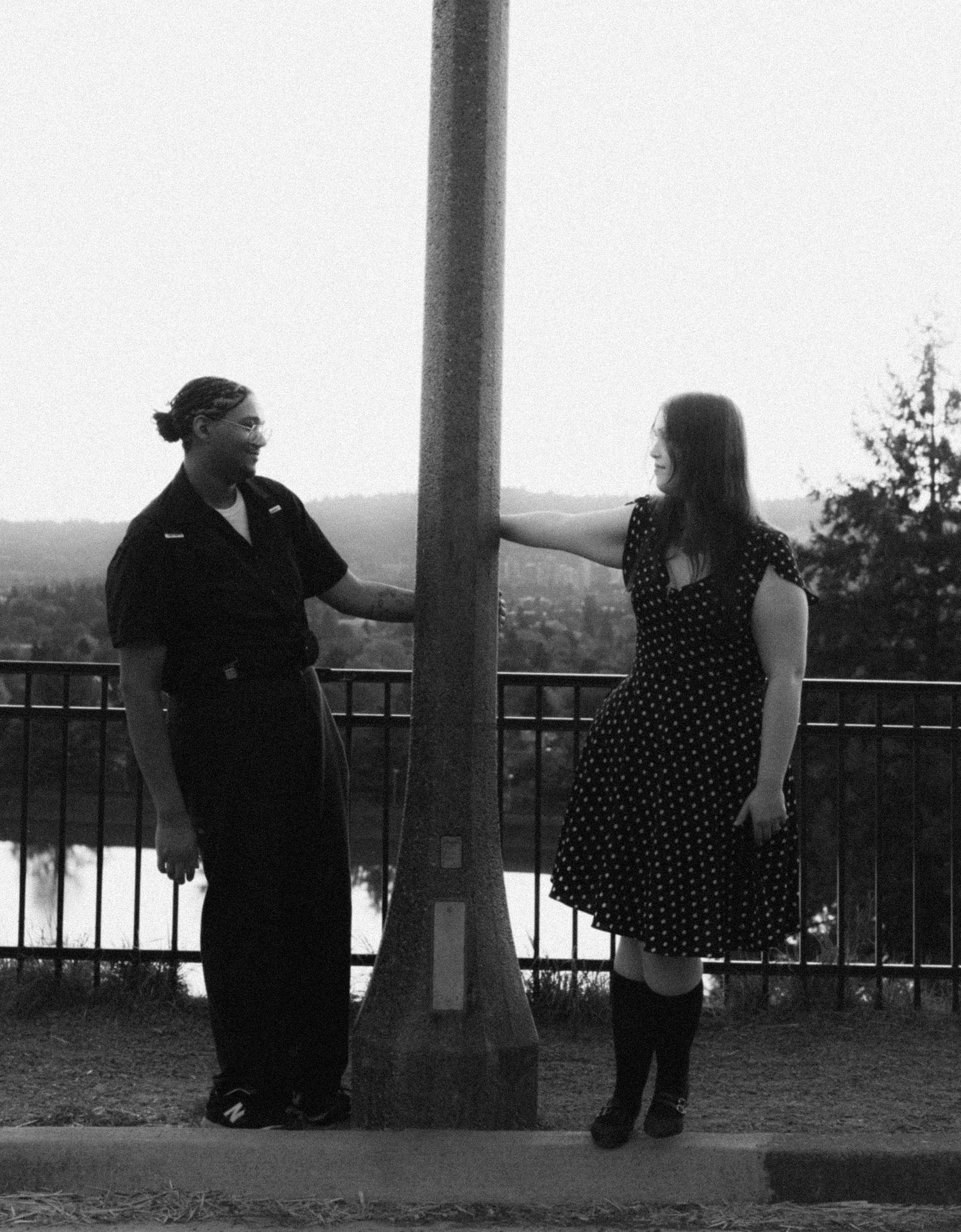 Black and white photo of a couple standing on opposite sides of a pole on a bridge, with trees in the background.