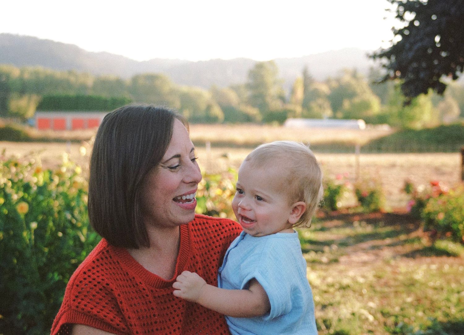 A smiling mother holds her happy blond toddler outdoors with golden sunlight and mountains in the background.