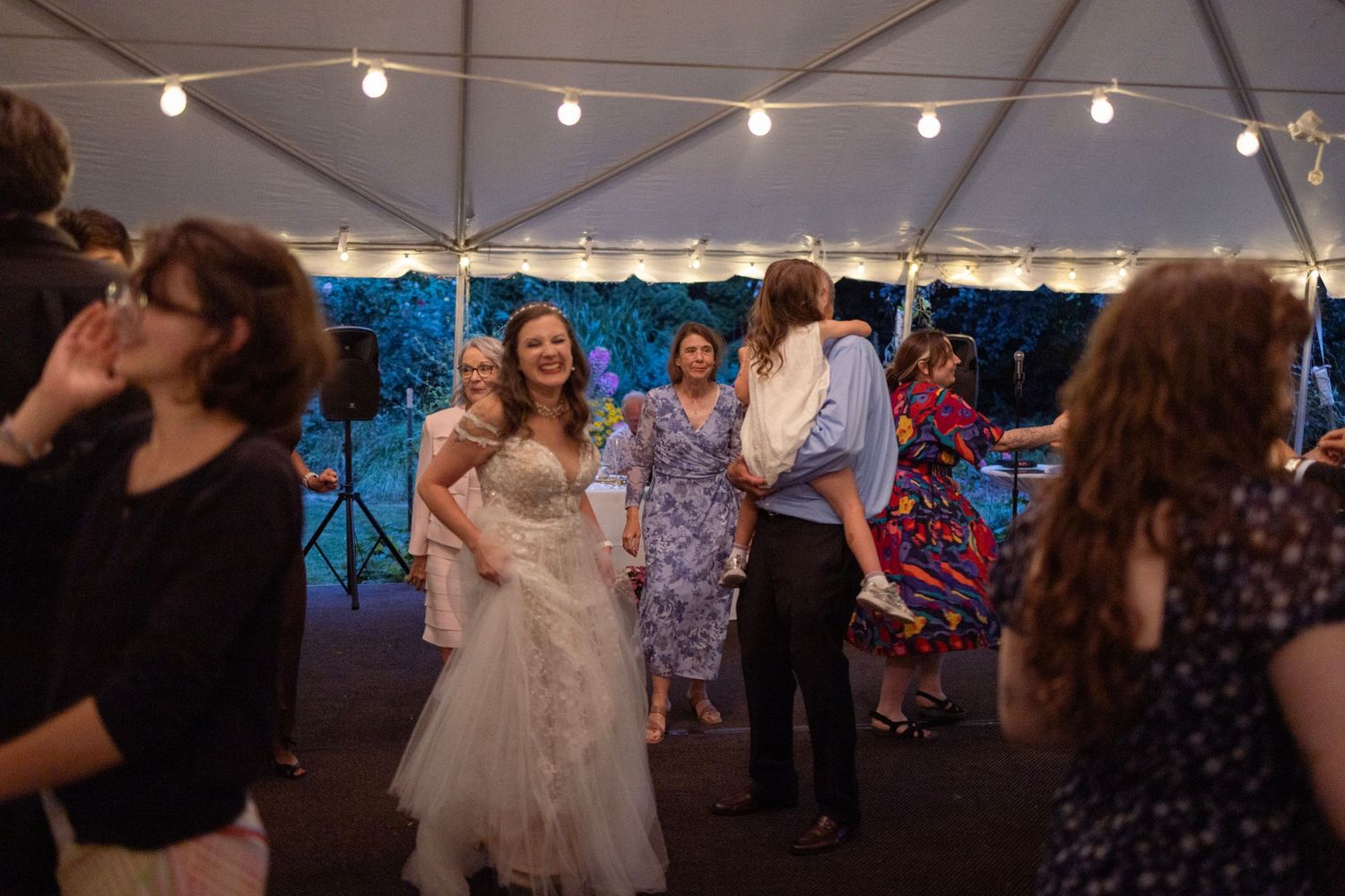 Bride in white lace gown smiling and dancing with guests under string lights at outdoor wedding reception tent.