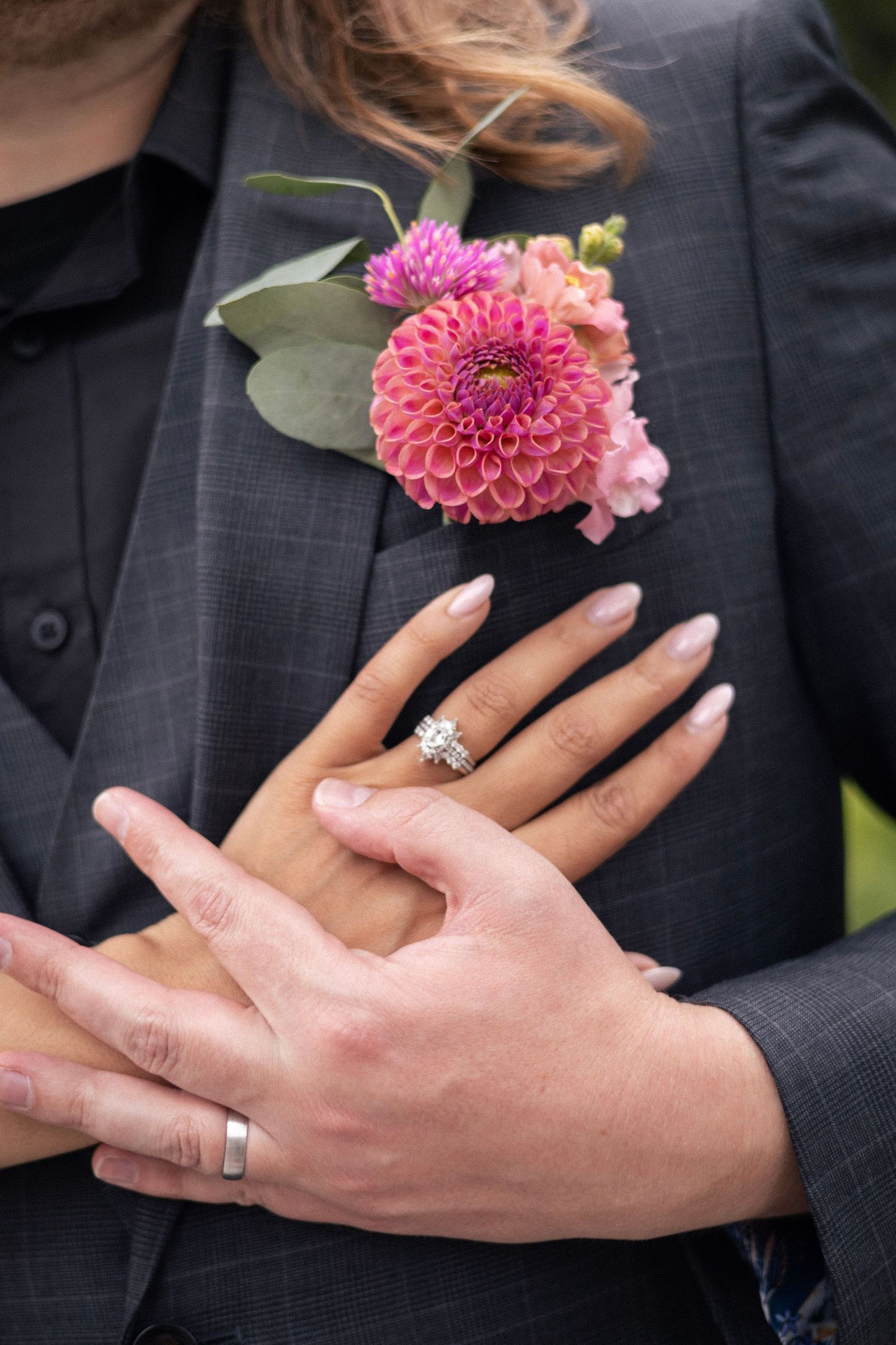 Couple showing wedding rings with pink dahlia boutonniere on groom's gray suit jacket.