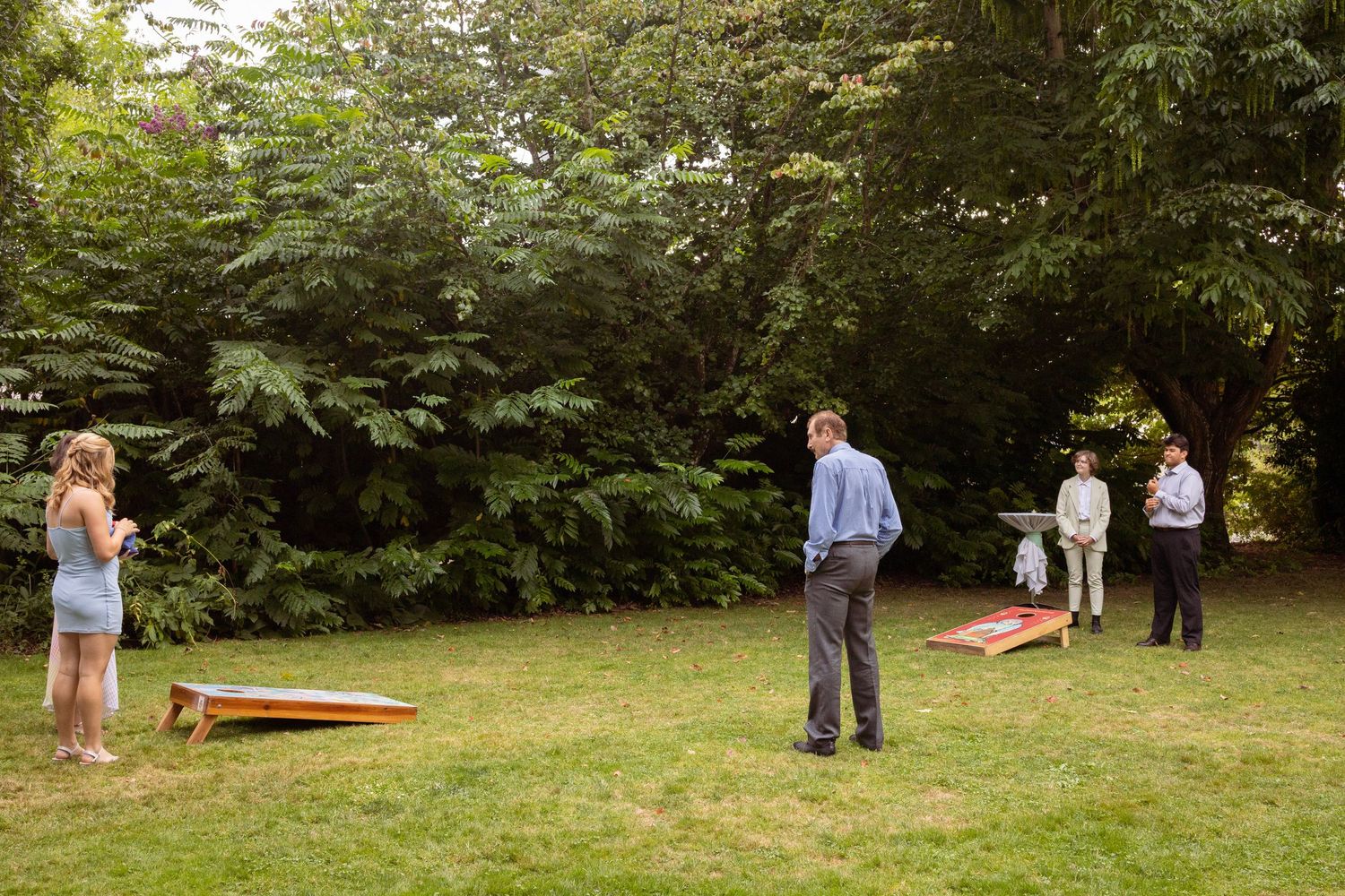 People playing cornhole lawn game outdoors on green grass surrounded by lush trees at a casual garden gathering.