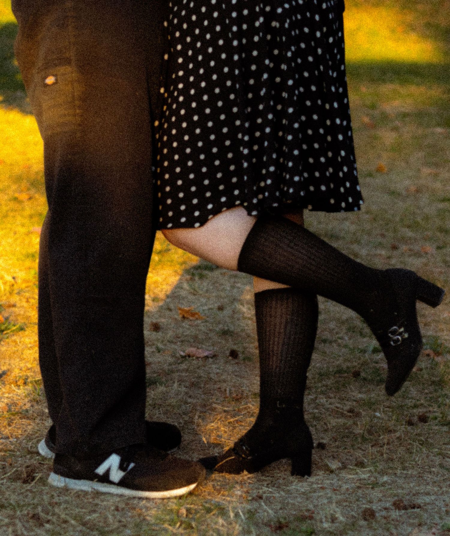Couple standing close together outdoors, woman in polka dot skirt with black heels, man in New Balance sneakers on grass.