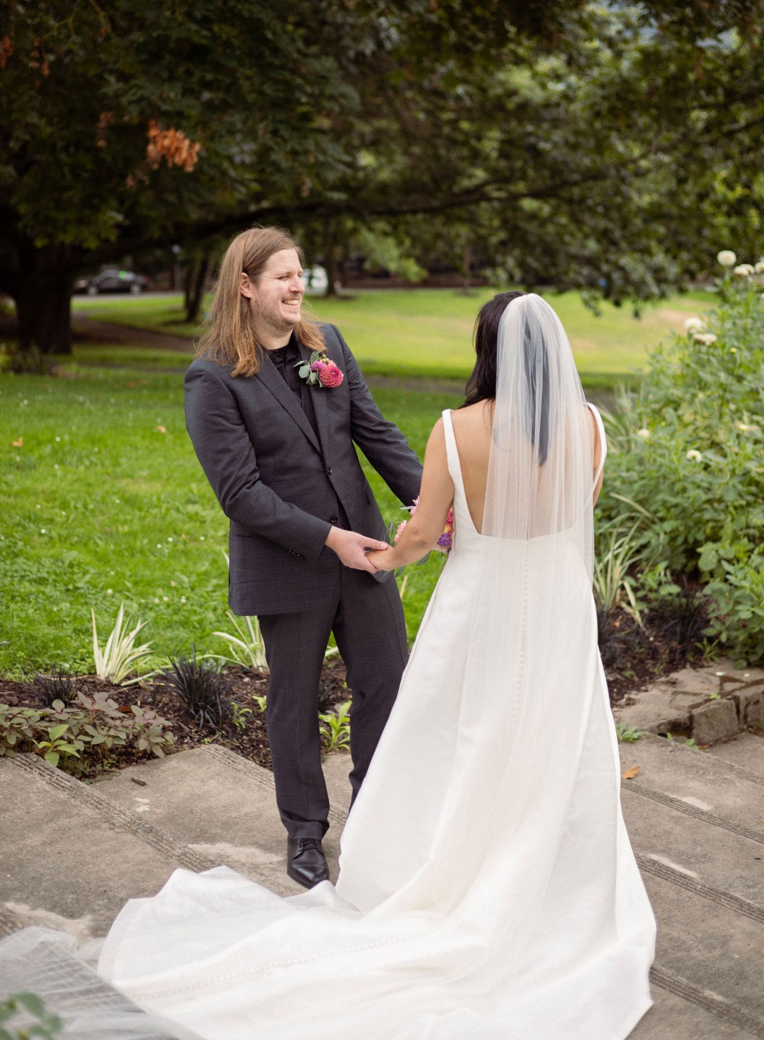 Bride in white gown and veil holds hands with smiling groom in dark suit outdoors.