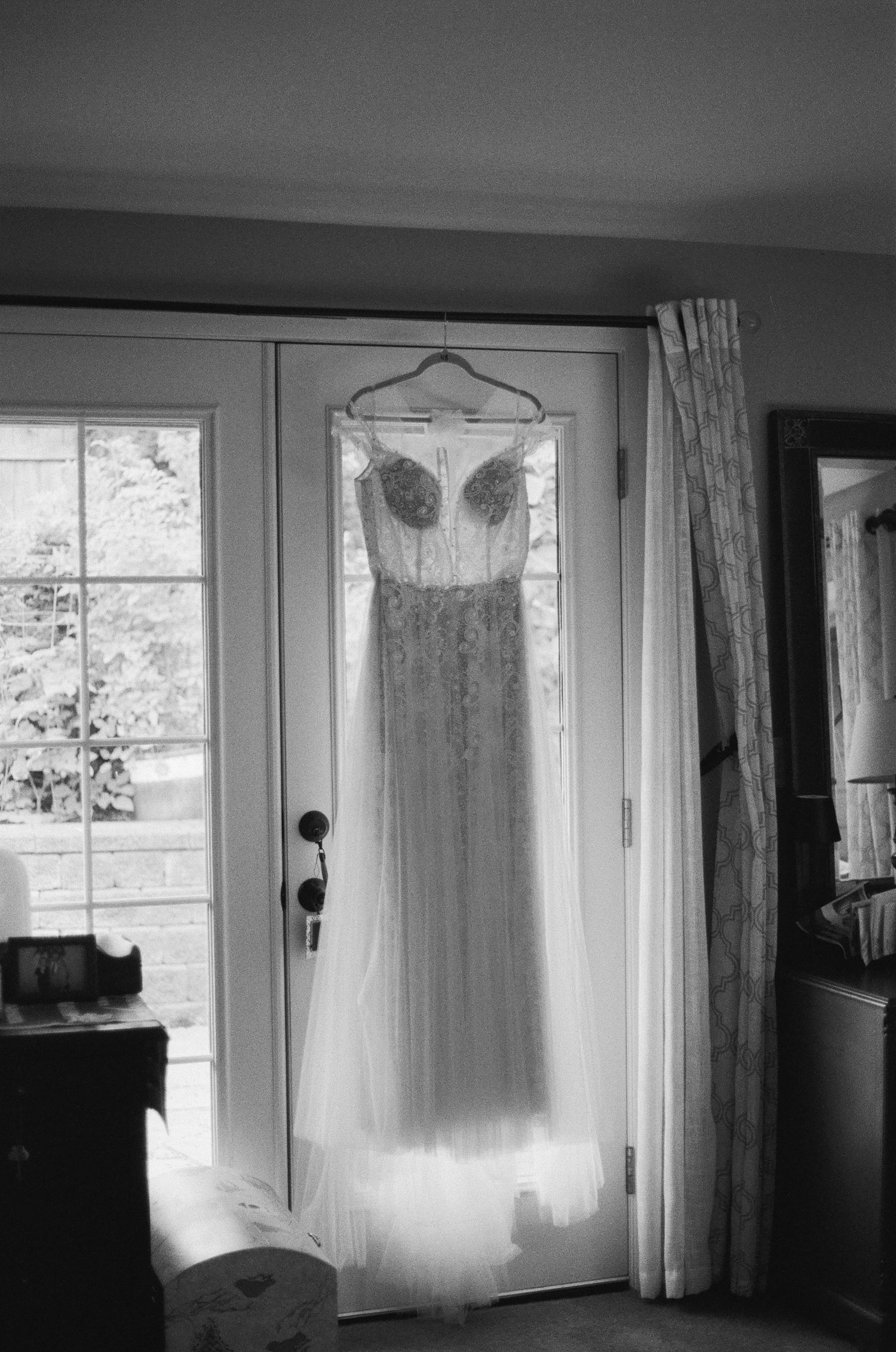 Black and white photo of an elegant lace wedding gown hanging on a door frame, backlit by natural window light.