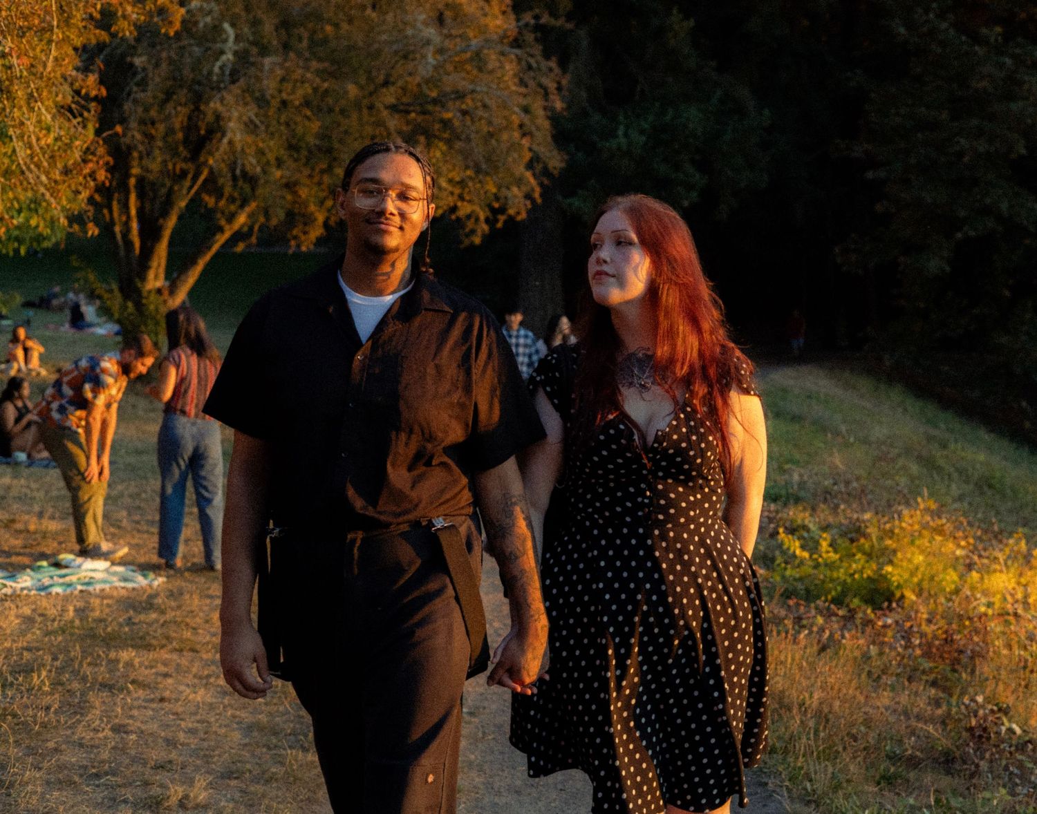 Couple holding hands walking on a sunlit park path, man in black shirt, woman in polka dot dress with red hair.