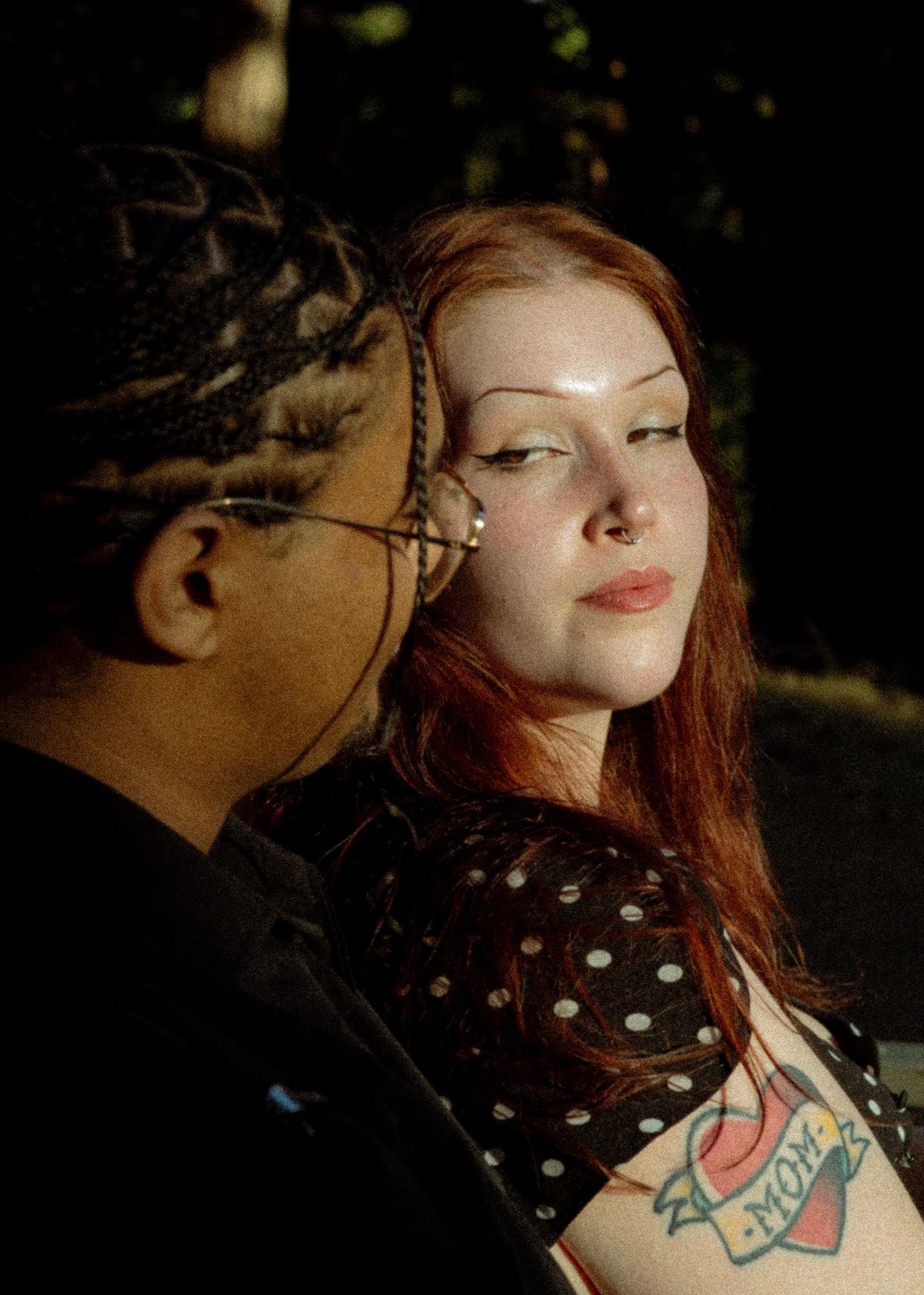 A redhead woman with a mom tattoo and nose ring sits beside a man with braids outdoors.