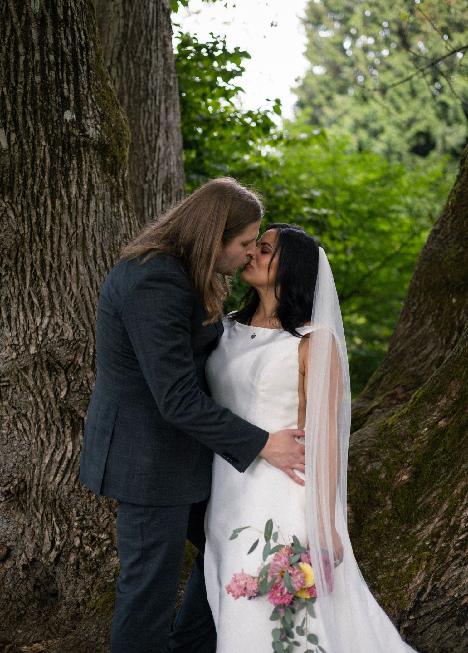 Bride and groom share a romantic kiss between two large trees, bride wearing white gown with veil and pink bouquet.