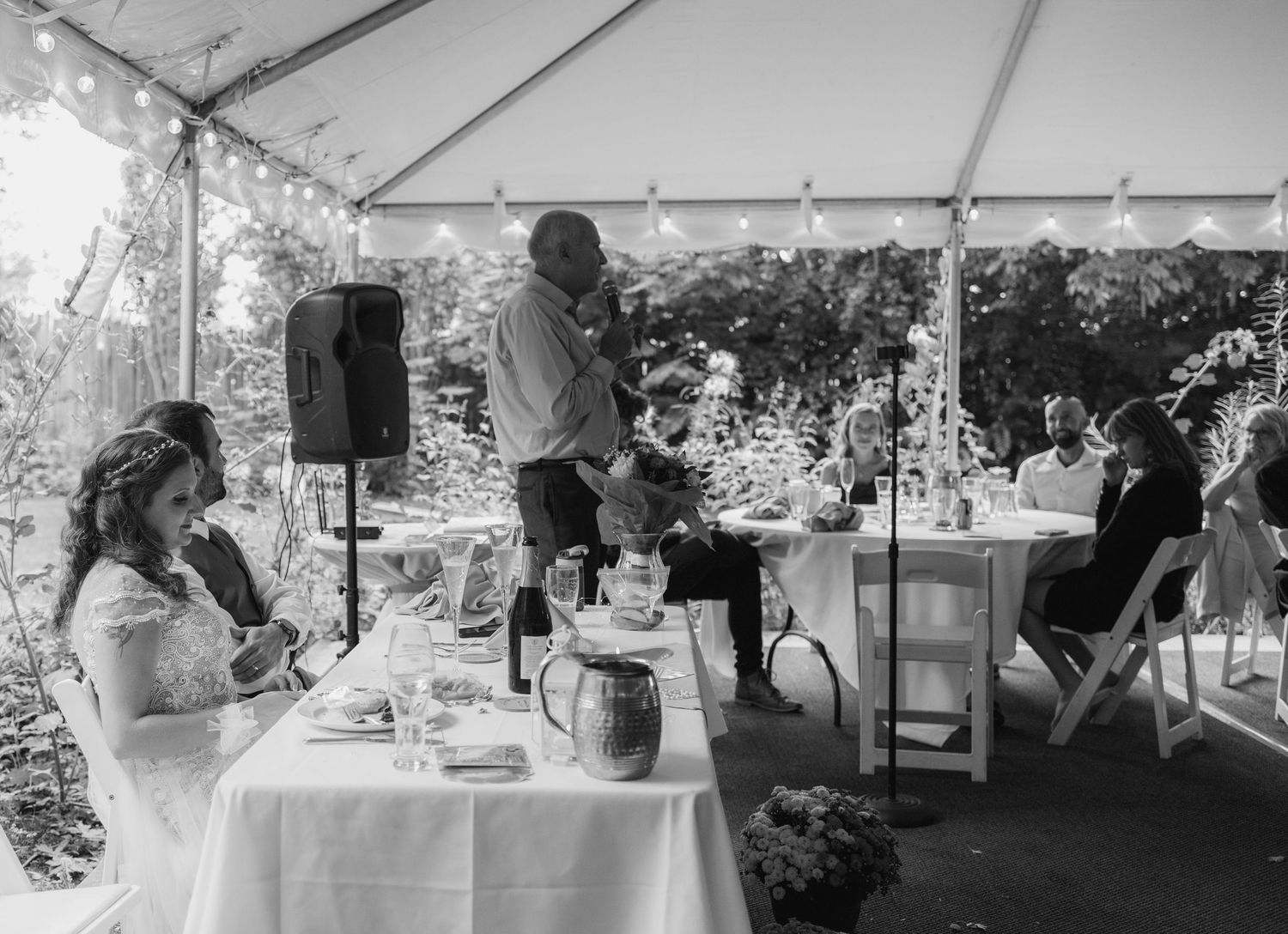 Black and white photo of wedding guests giving toast at outdoor reception under tent.