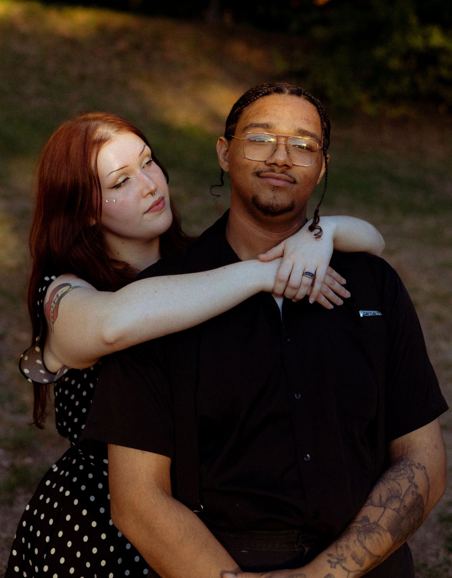 A couple embraces outdoors, woman with red hair in polka dot dress rests arm on tattooed man in black shirt.