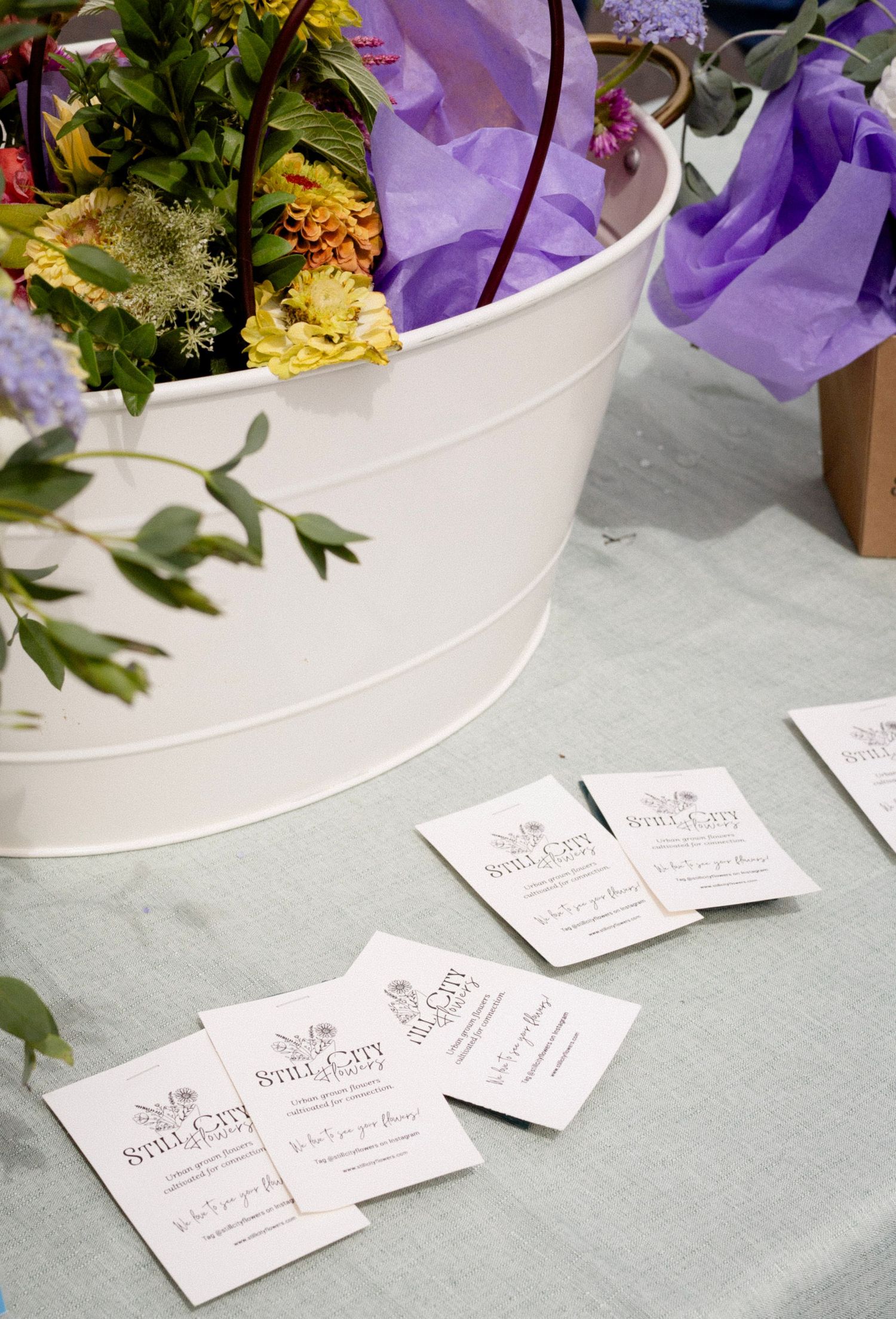 White bucket with colorful flowers and Still City business cards displayed on a light blue tablecloth.
